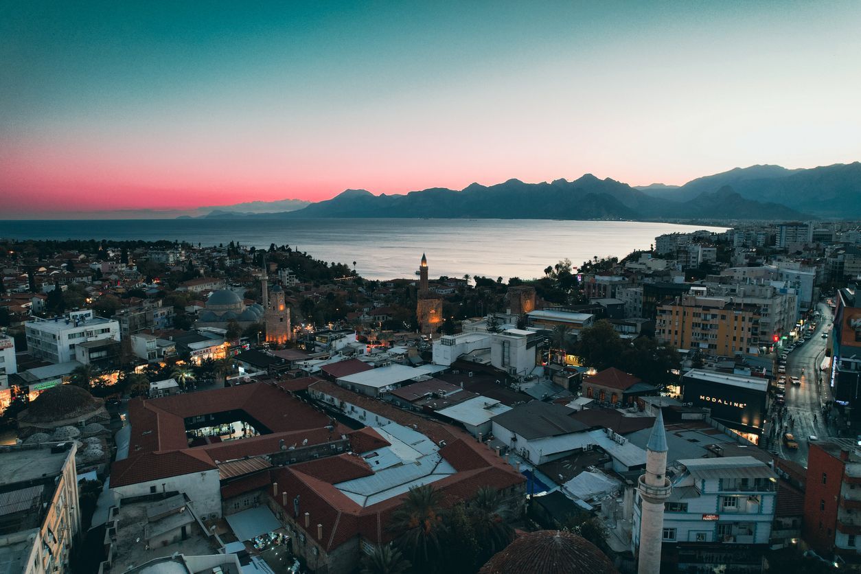 Aerial view of a coastal city with minarets overlooking a bay at sunset, with mountains in the distance.