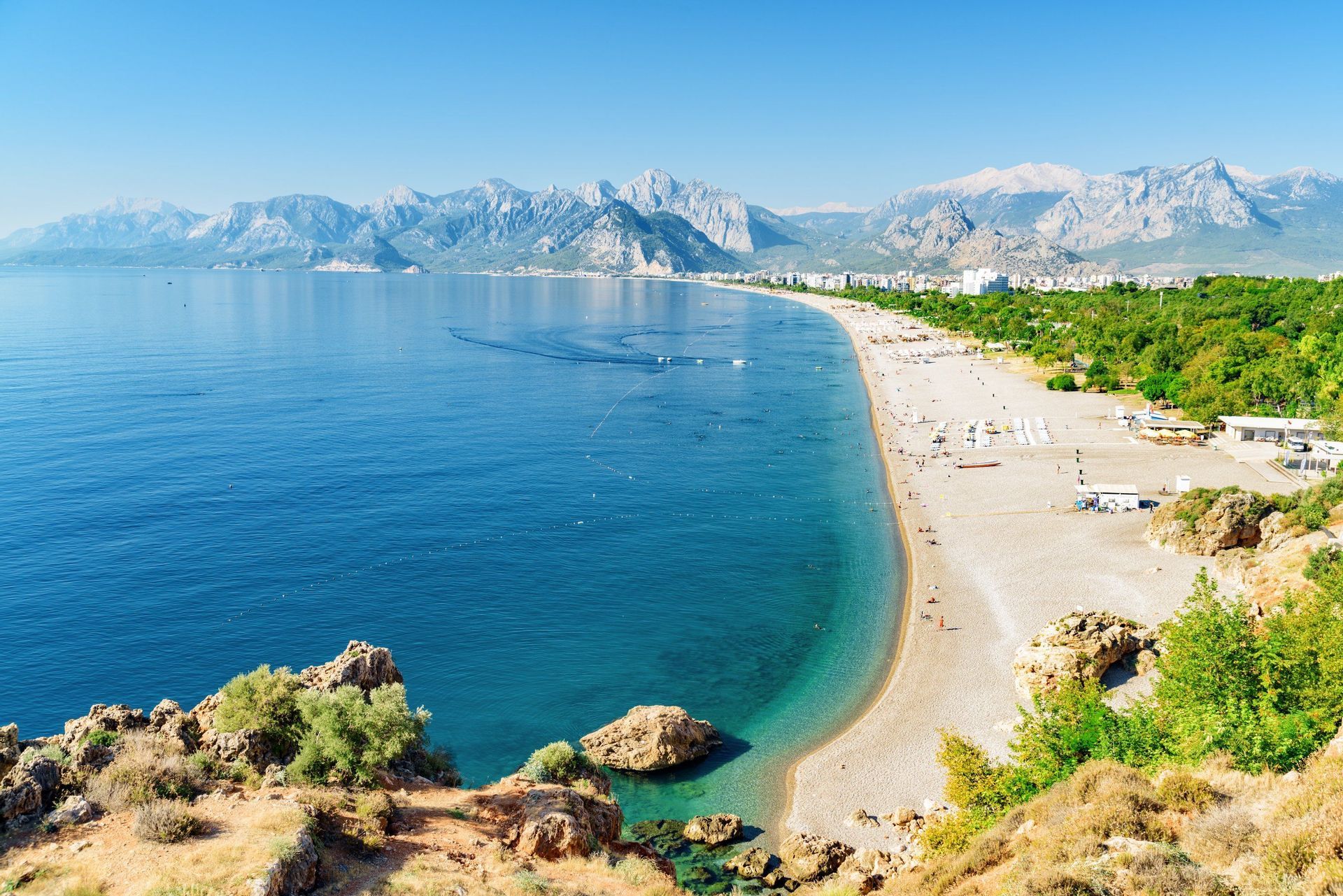 An aerial view of a long, curving beach with turquoise water, a coastal city, and a mountain range in the background under a clear sky.