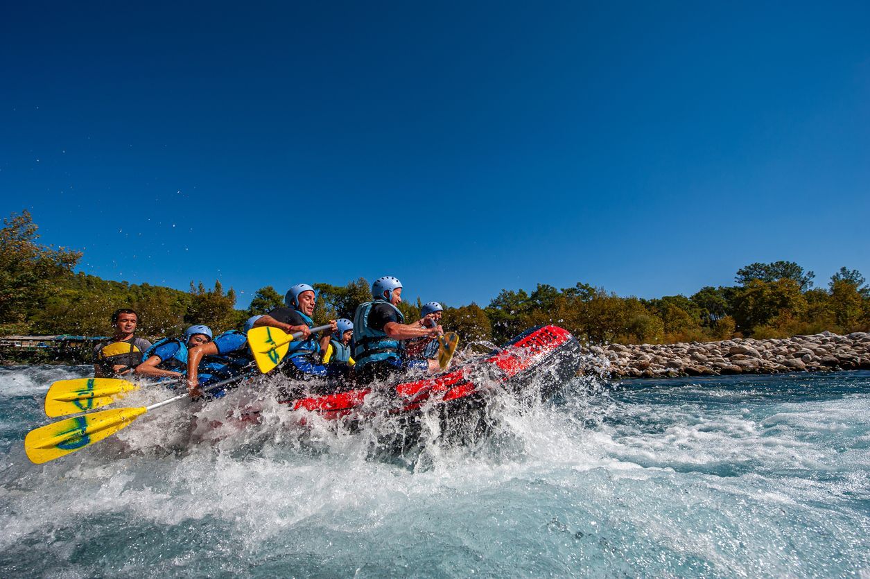 A WeRoad group trip paddles a red raft through splashing whitewater rapids under a clear blue sky.
