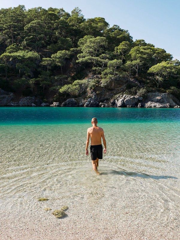 A man in black shorts, seen from behind, wades into shallow, clear turquoise water towards a tree-covered shoreline.