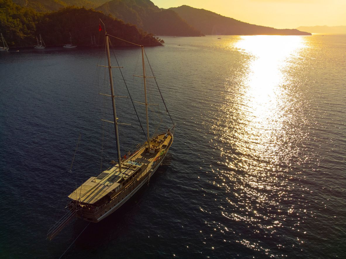 An aerial view of a wooden gulet sailboat anchored in a calm bay at sunset, with golden light reflecting on the water.