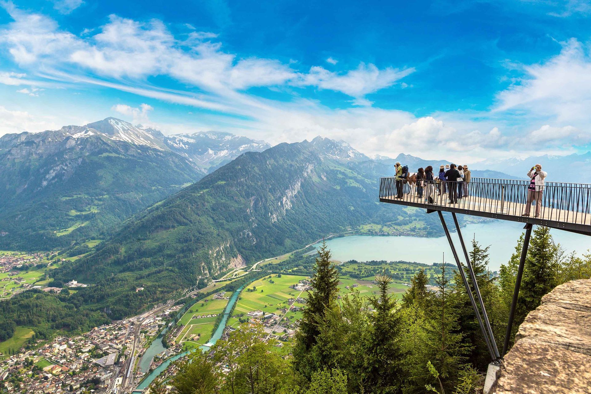 Un gruppo WeRoad ammira una vista panoramica mozzafiato su una valle montana, un lago e una città da una piattaforma sospesa.