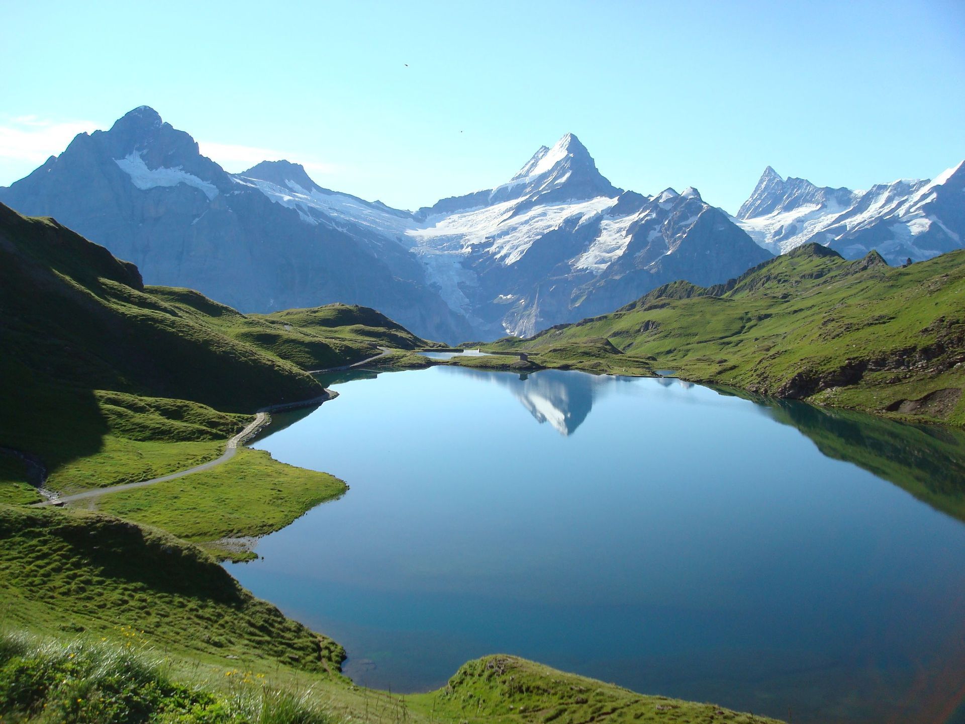 A still alpine lake reflects a snow-capped mountain peak, surrounded by green hills under a clear blue sky.