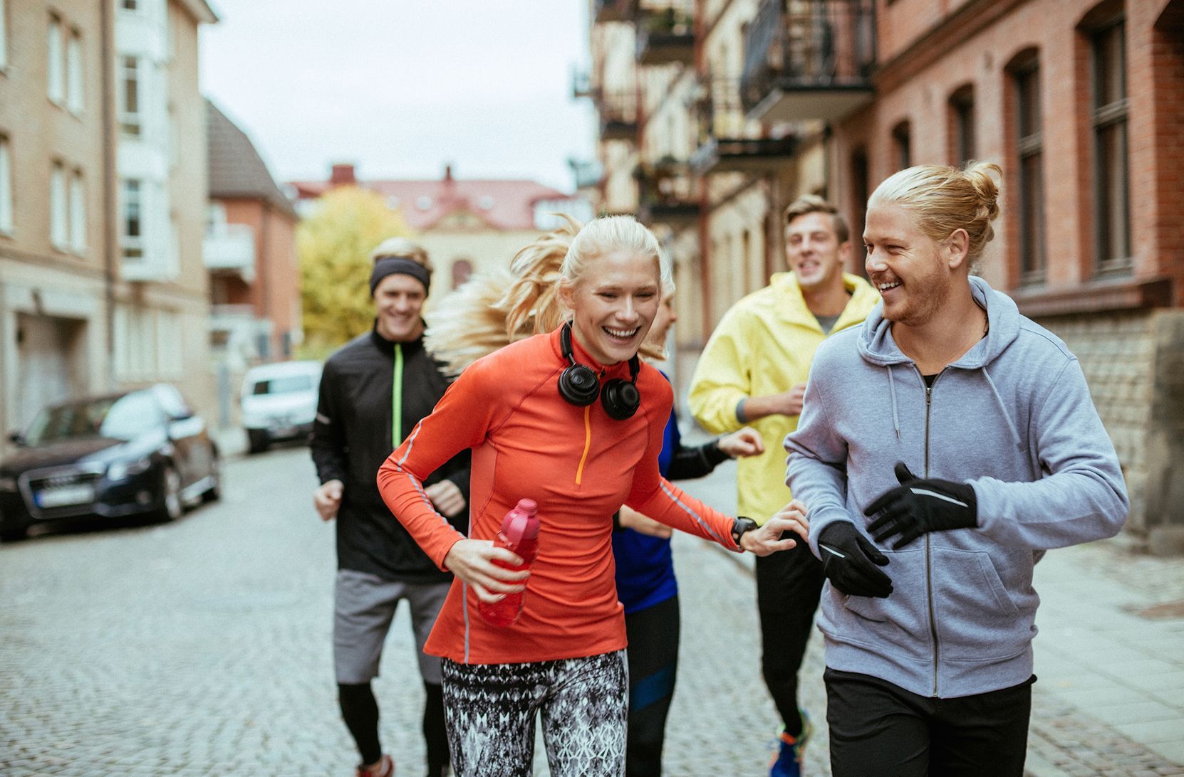 Un gruppo di amici in abbigliamento sportivo sorride mentre fa jogging insieme su una strada acciottolata in città.