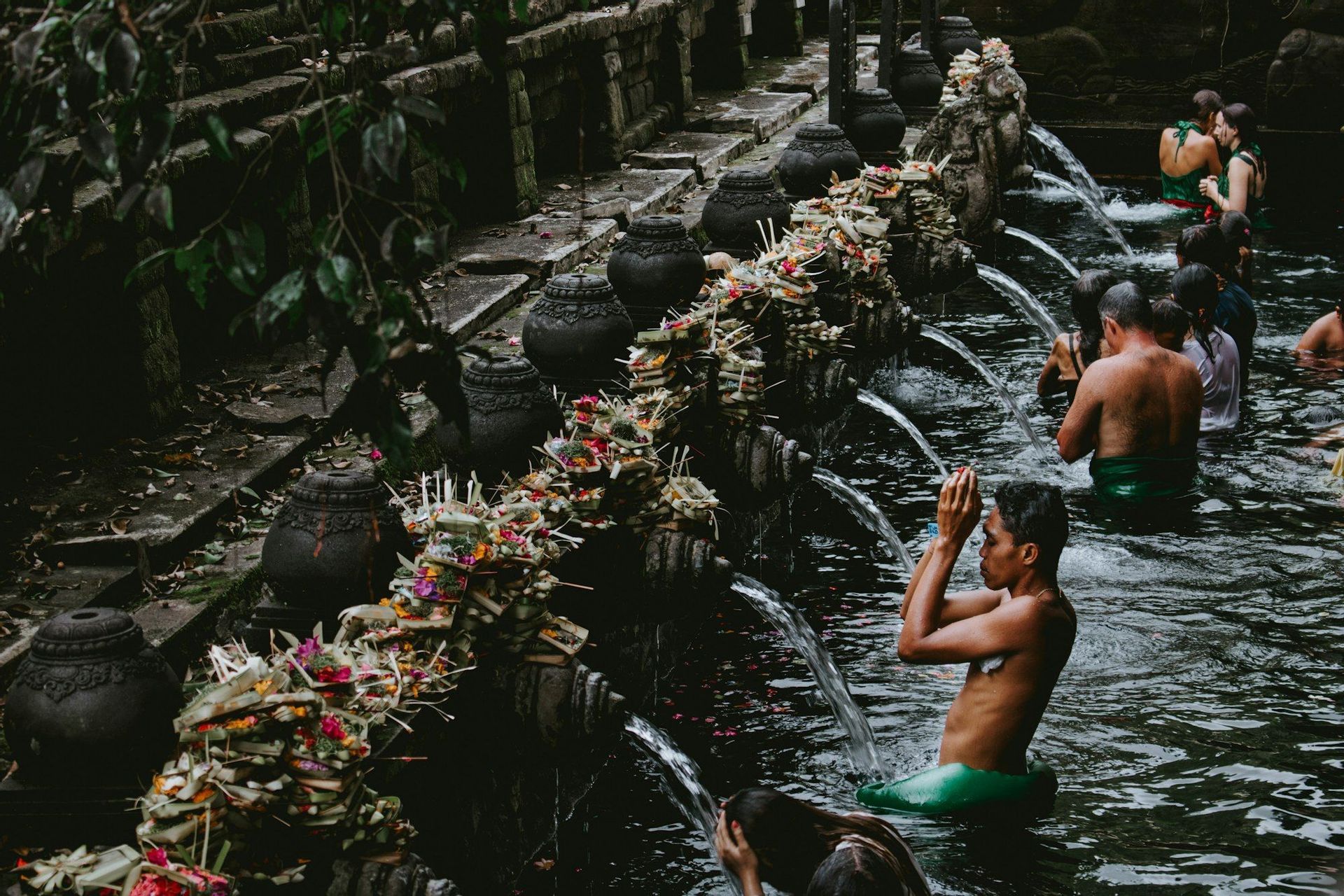Eine Gruppe von Menschen nimmt an einem Reinigungsritual teil und badet in einer heiligen Quelle unter Steinausgüssen, die mit Opfergaben geschmückt sind.