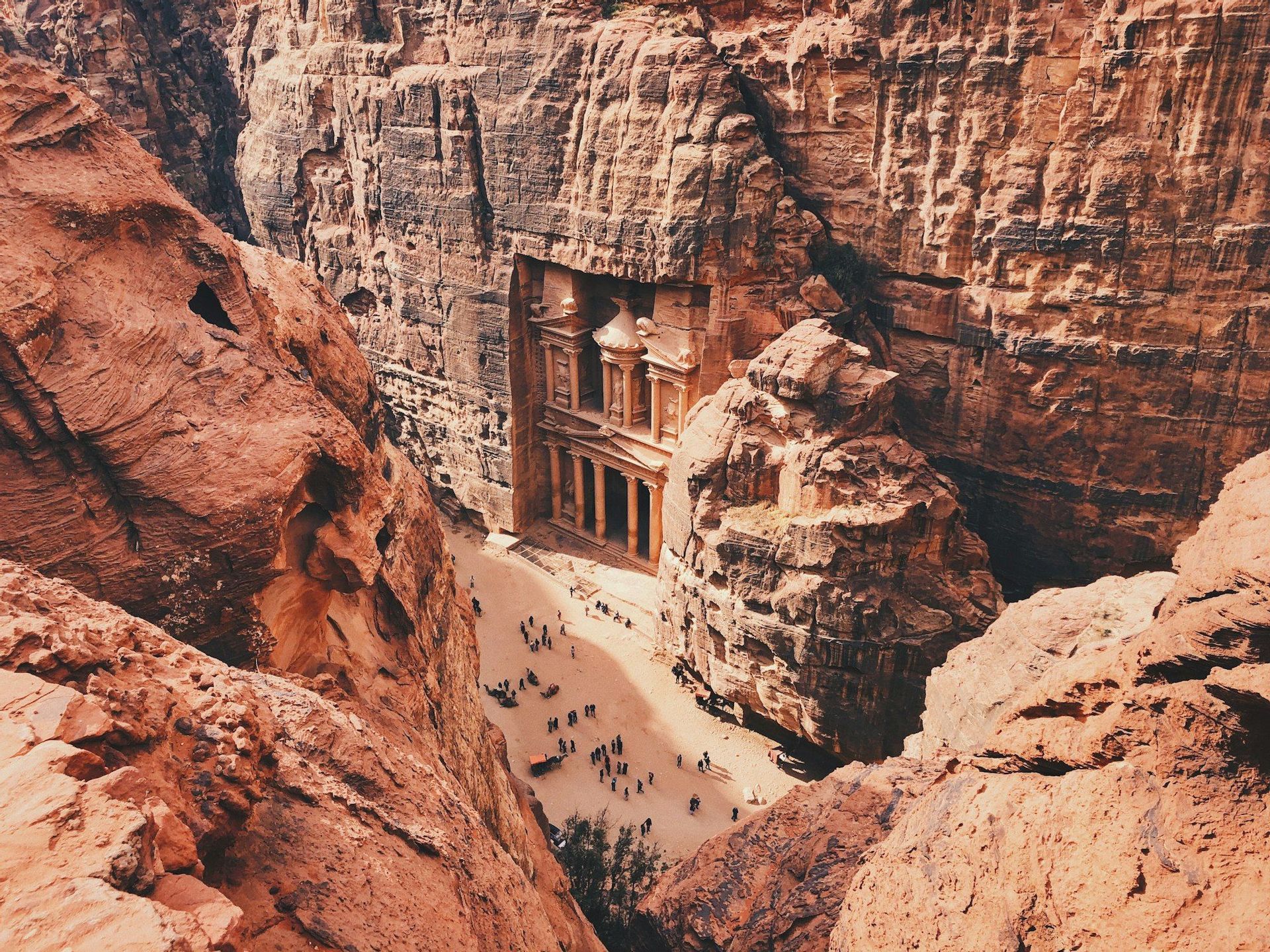 Ein erhöhter Blick auf einen großen Tempel, der in die rote Felswand einer Schlucht gehauen ist, mit Menschenmengen, die sich im sonnenbeschienenen Bereich darunter versammelt haben.