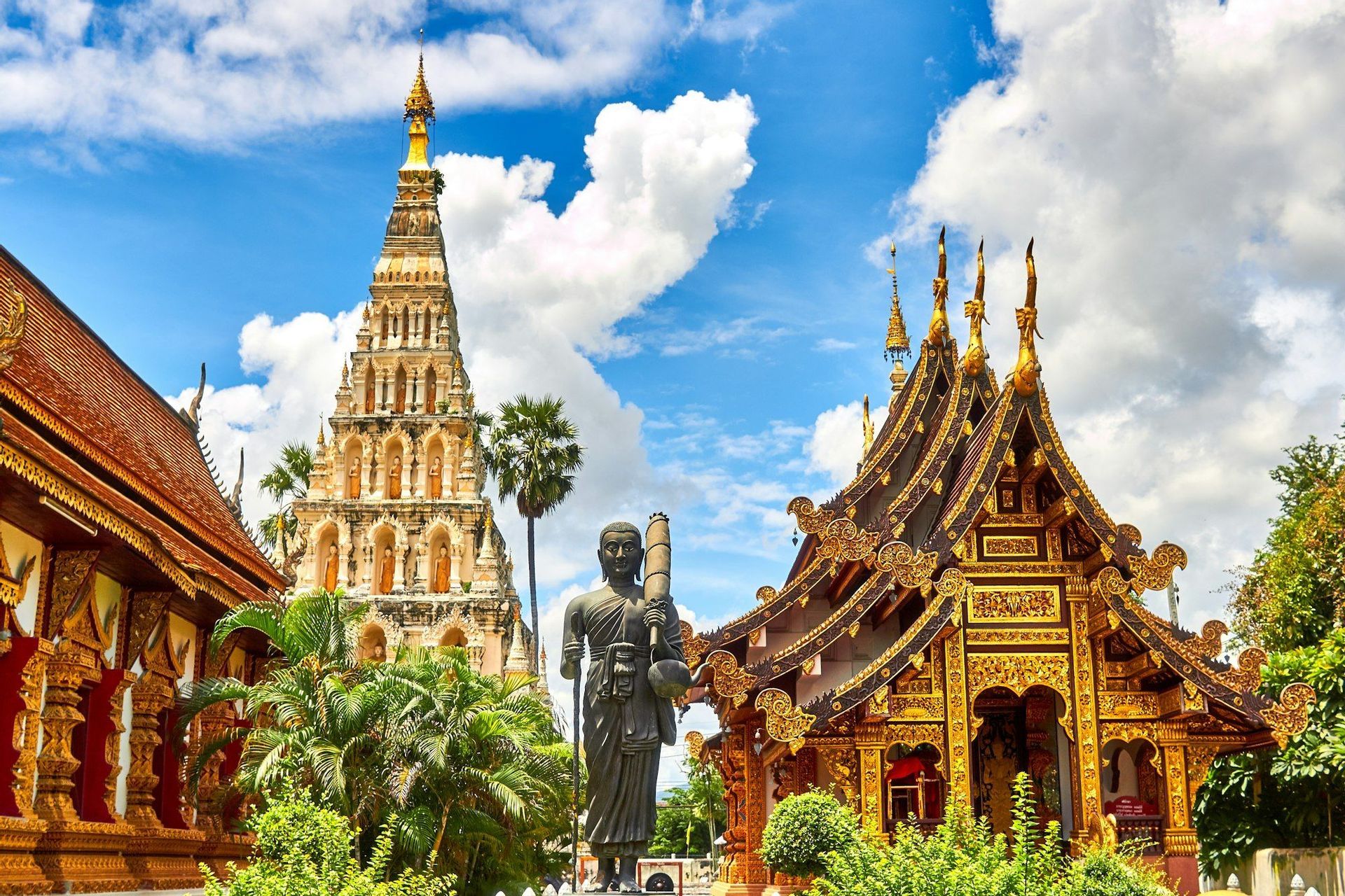 Ein kunstvoll verzierter goldener Tempel und eine hohe steinerne Stupa stehen neben einer Mönchsstatue, umgeben von Grün unter blauem Himmel.