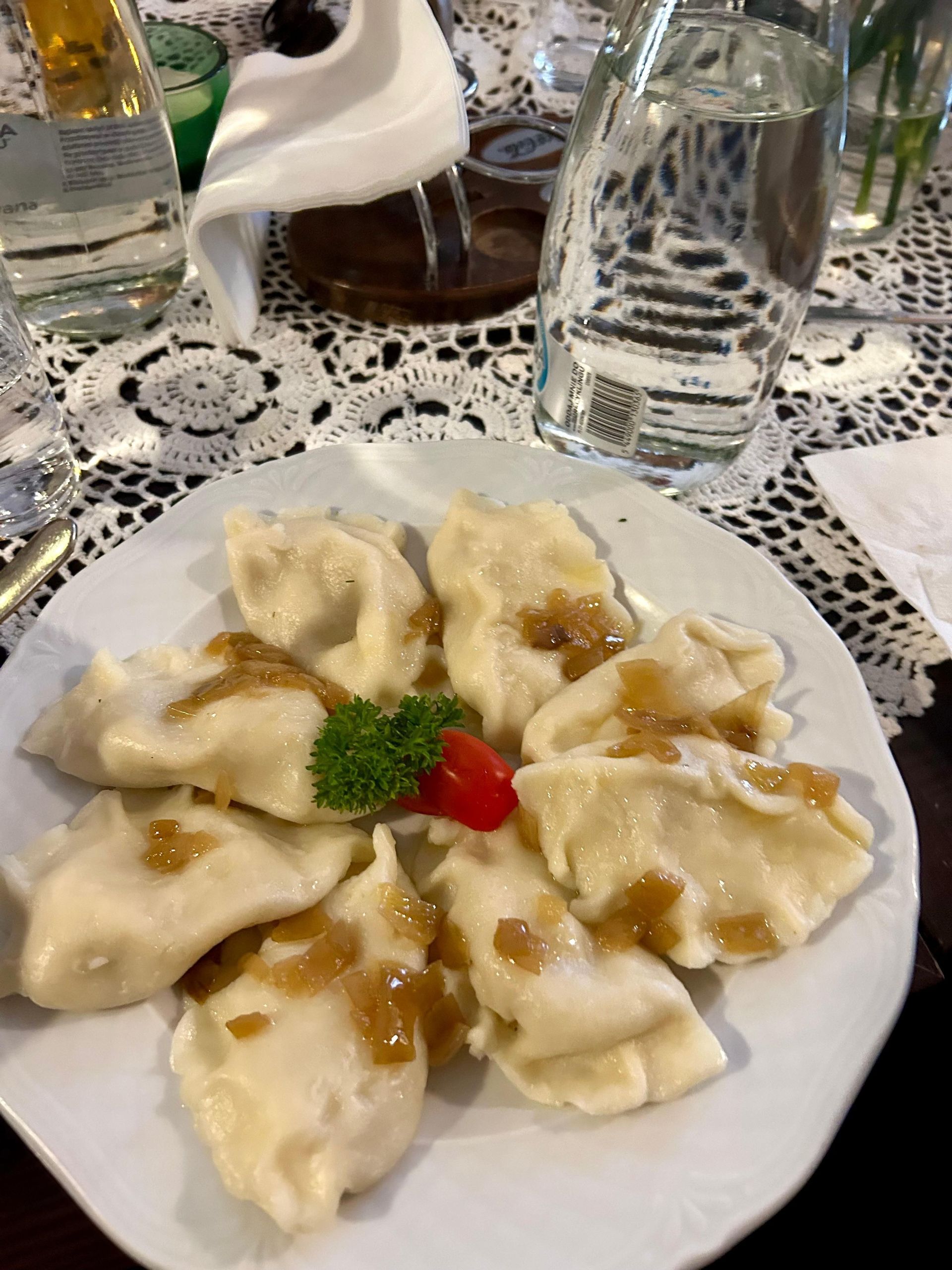 A white plate of pierogi dumplings topped with fried onions, garnished with parsley and tomato, sits on a lace doily.