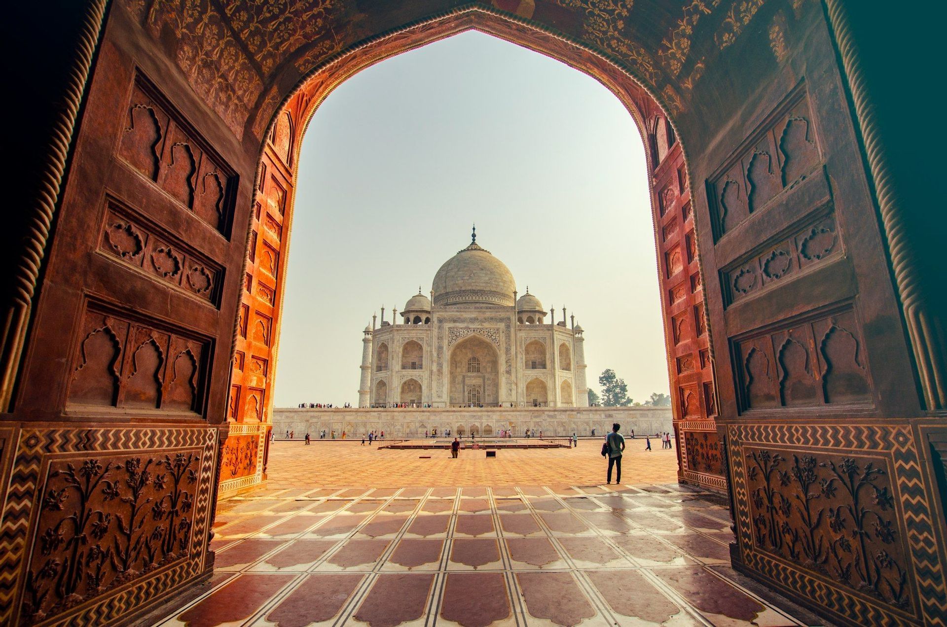 The Taj Mahal is viewed from inside a large, ornate arched gateway with open wooden doors, framing the white marble monument.