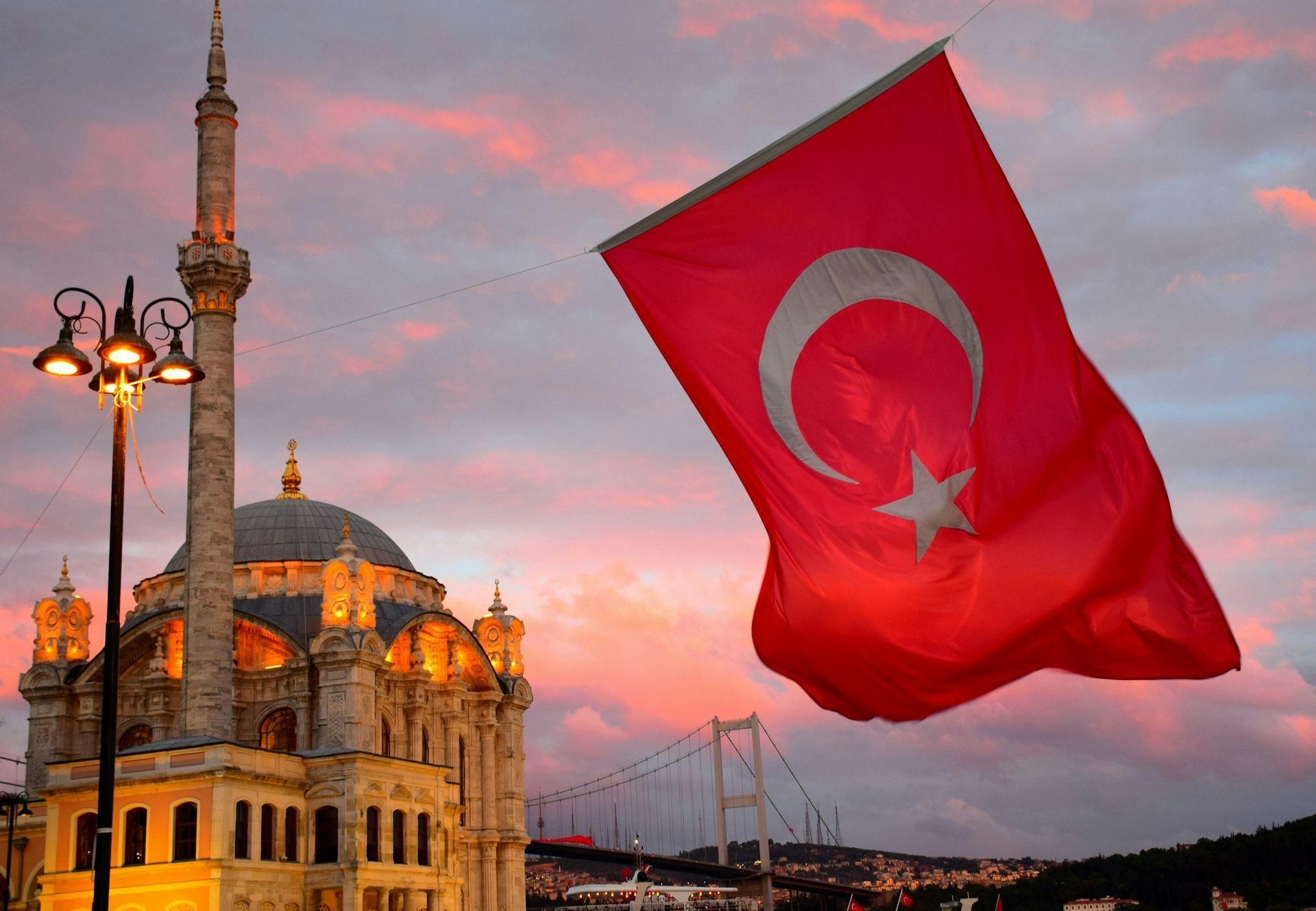 The Turkish flag waves in front of an illuminated mosque and a large suspension bridge against a colorful sunset sky.