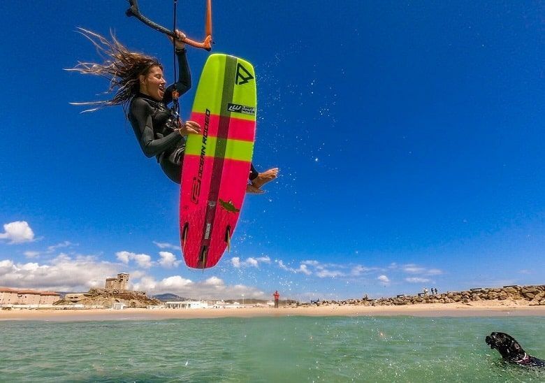 Una mujer en traje de neopreno vuela con su kiteboard sobre el mar en un día soleado, con una playa y un perro de fondo.