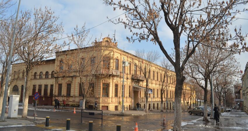 Un grande edificio storico giallo pallido all'angolo di una strada, con alberi spogli e persone che camminano sul marciapiede bagnato.