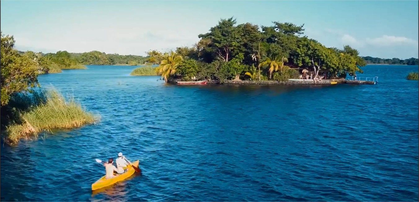 Two people in a yellow kayak paddling across a blue lake towards a small, tree-covered island.