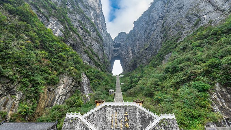 Una lunga scalinata ripida sale su una montagna ricoperta di verde verso un imponente arco di roccia naturale situato tra due scogliere.