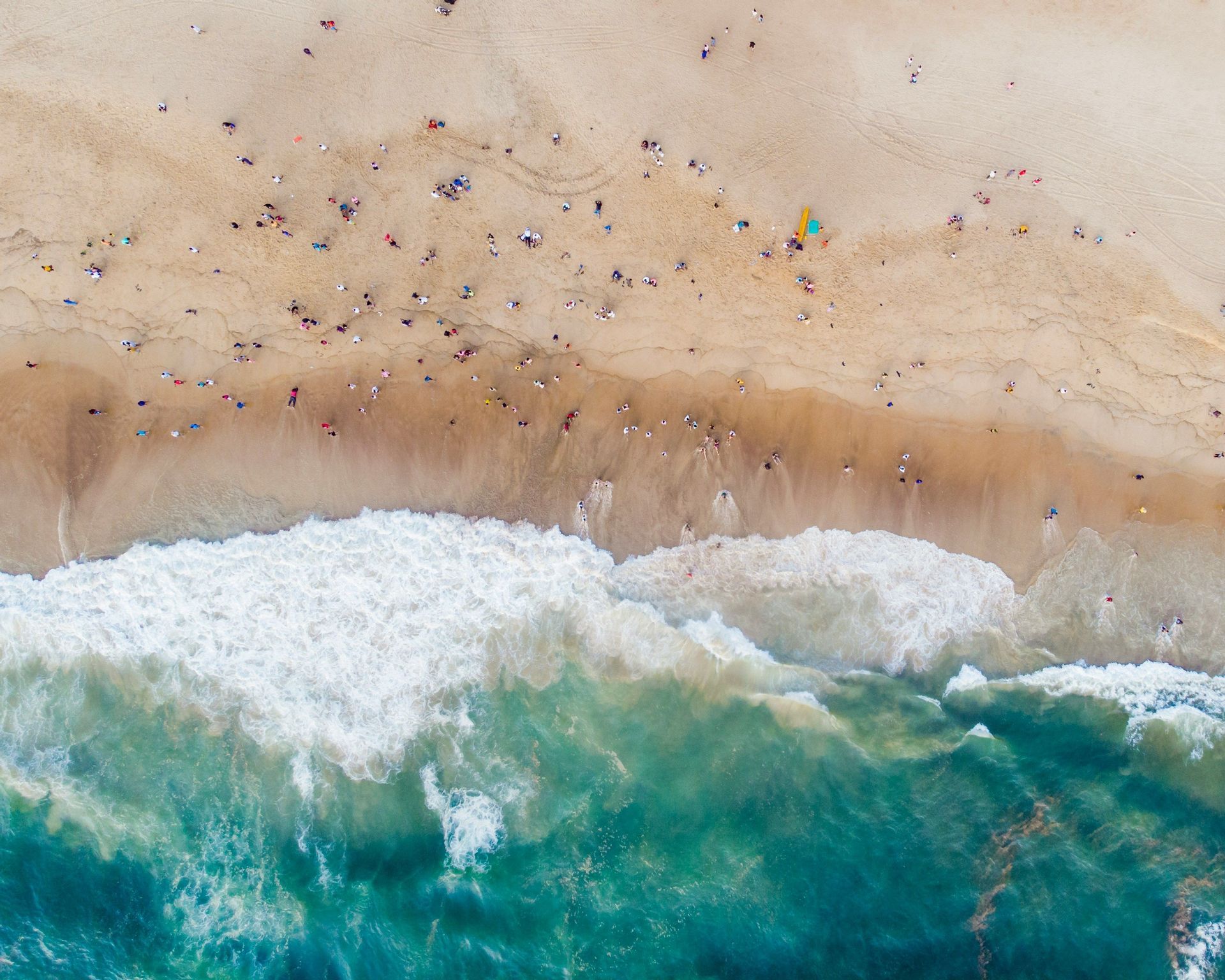 Vista aerea dall'alto di una spiaggia sabbiosa affollata con persone sparse lungo la riva mentre onde turchesi si infrangono.