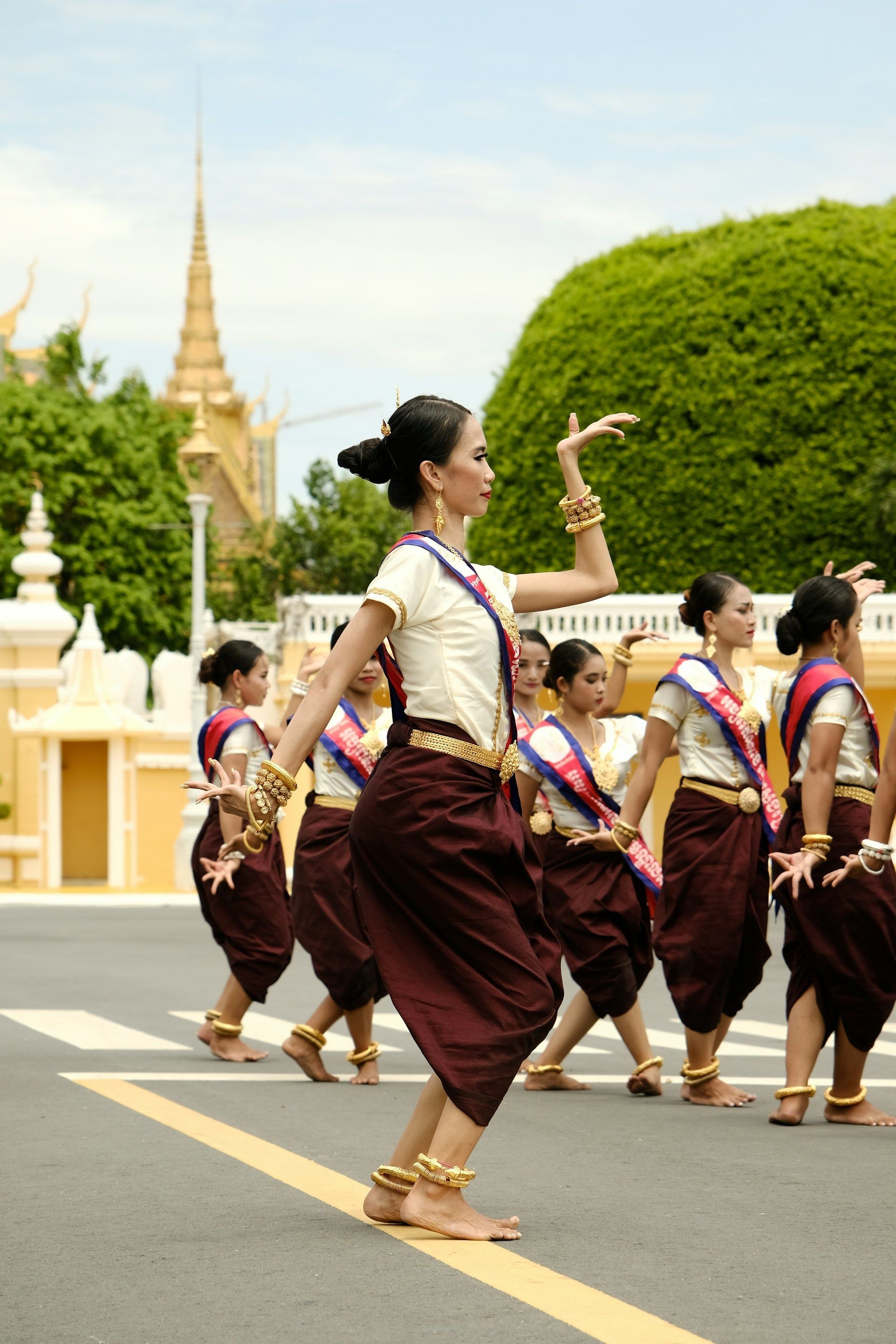 Un grupo de mujeres con atuendos tradicionales de color granate y crema, y joyas de oro, realizan una danza cultural en la calle.