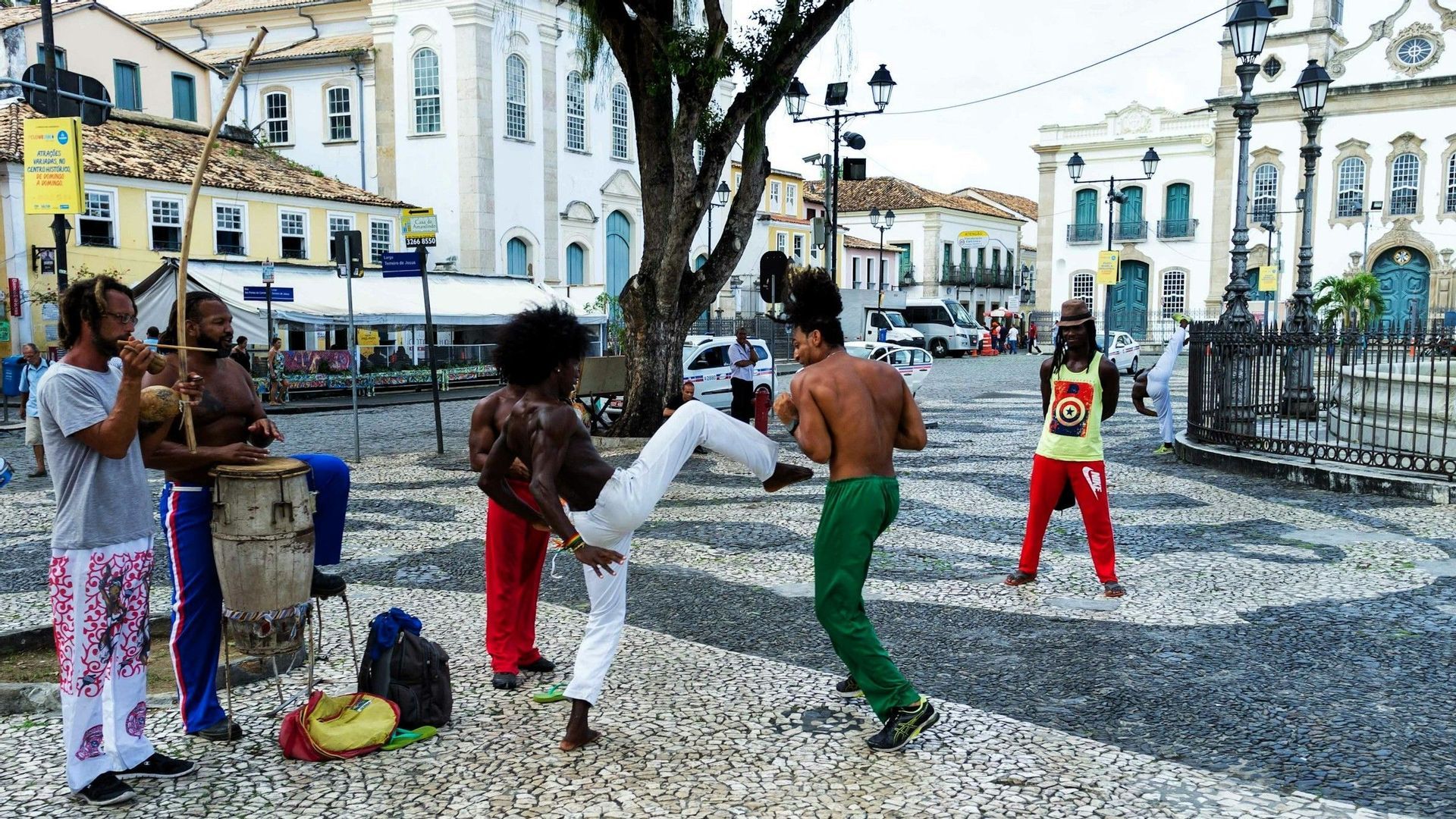 Eine Gruppe von Männern führt Capoeira, eine brasilianische Kampfkunst, auf einem Kopfsteinpflasterplatz auf, der von historischen Gebäuden umgeben ist.