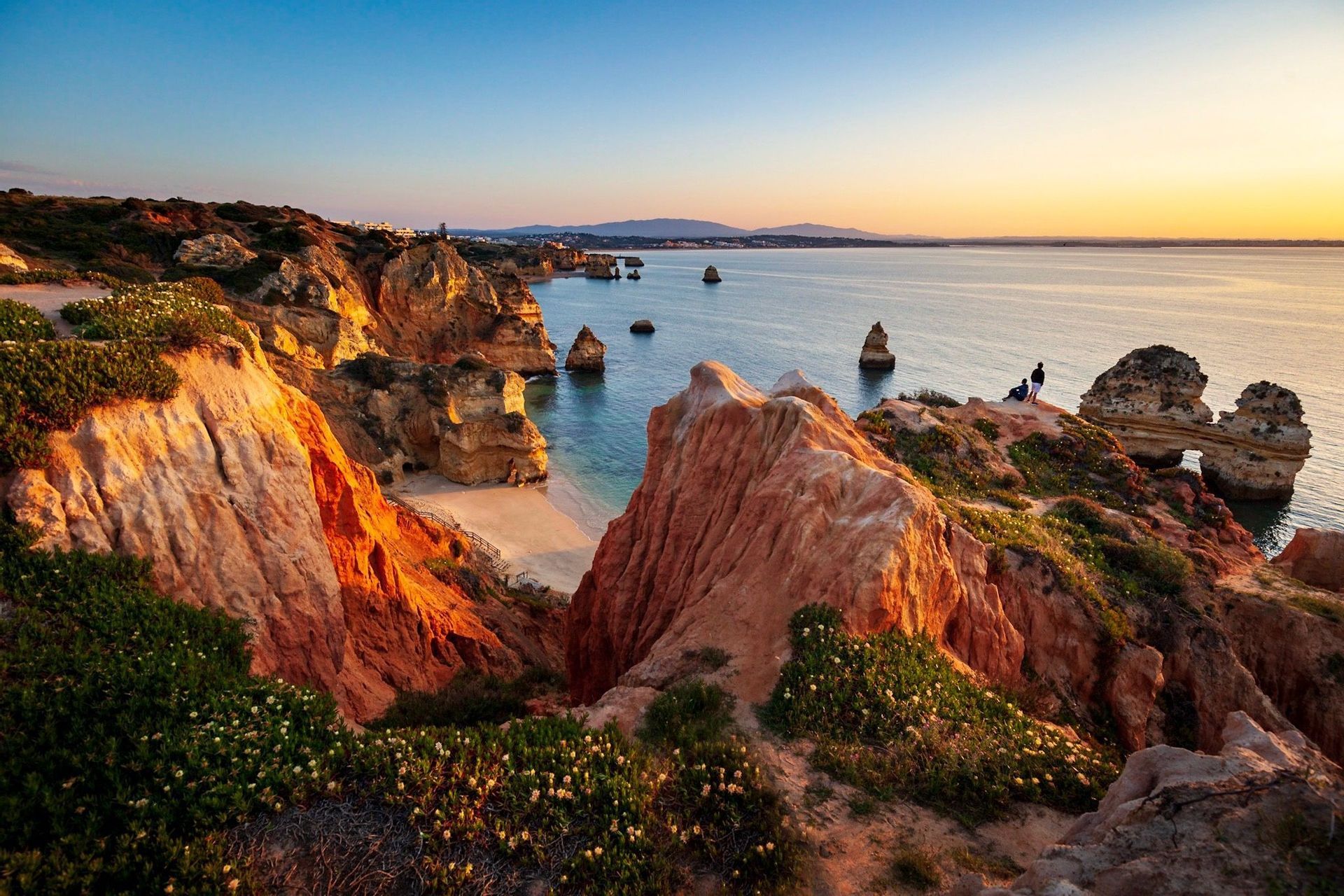 Rugged red cliffs covered in green vegetation overlook a calm sea with sea stacks and a sandy cove at sunset.