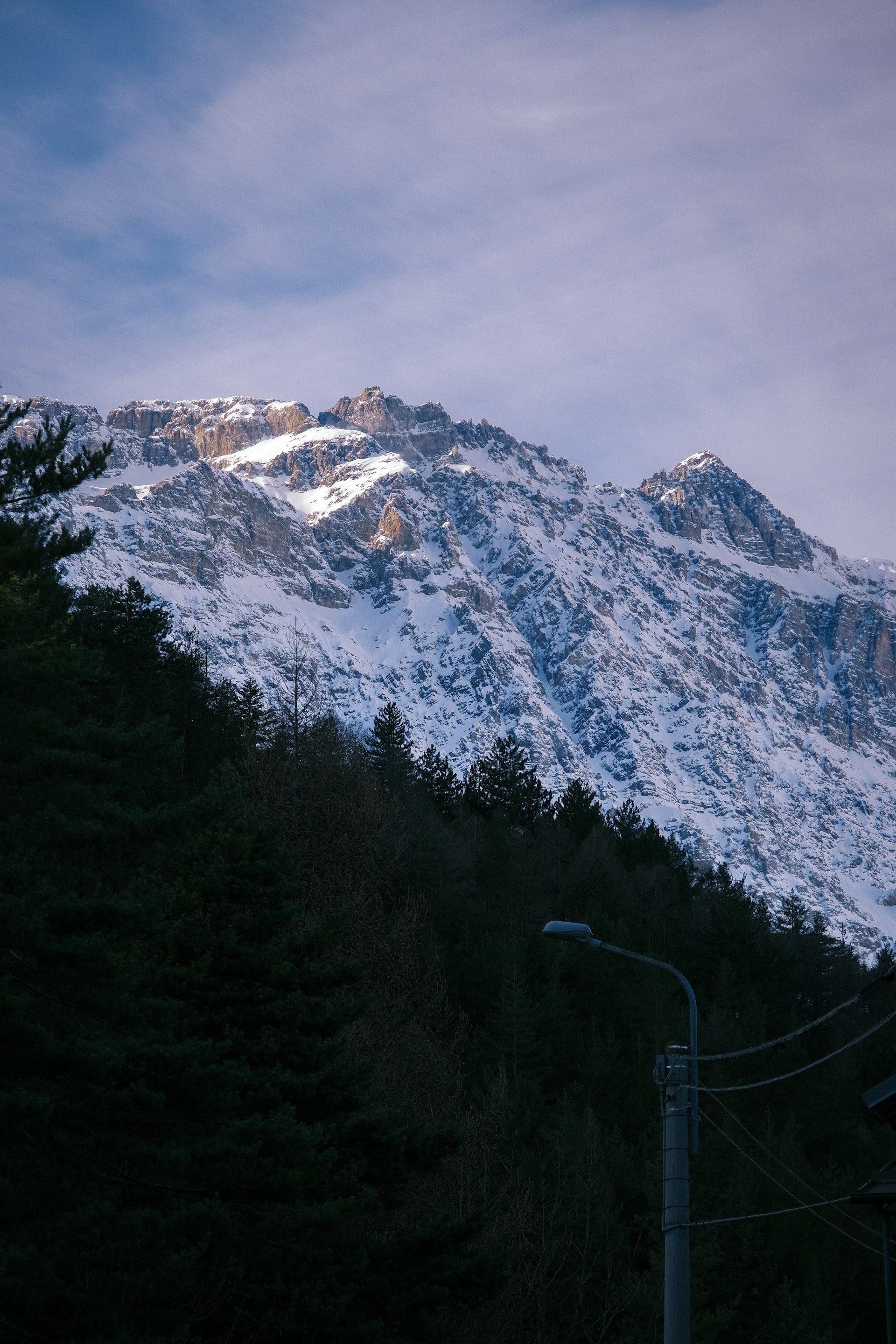 La luce del sole illumina le cime di una grande montagna innevata che sovrasta una fitta e scura foresta di pini sotto un cielo blu.