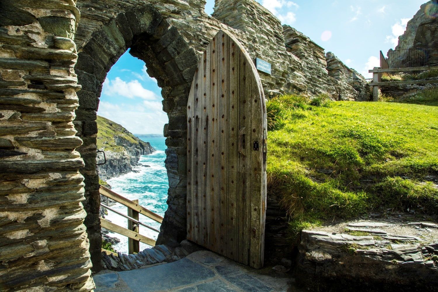 Una porta di legno aperta in un arco di pietra rivela una vista dell'oceano turchese che si infrange contro una costa rocciosa.