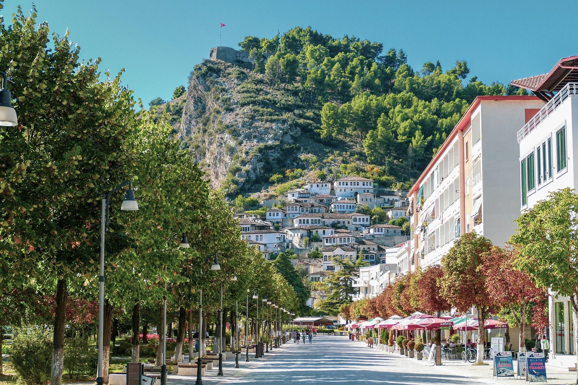 Une rue piétonne ensoleillée, bordée d'arbres, menant à un village perché aux maisons blanches et à une forteresse sur son sommet.