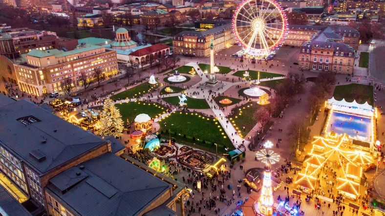 An aerial view of a bustling city square at dusk, filled with a Christmas market, an illuminated Ferris wheel, and an ice rink.
