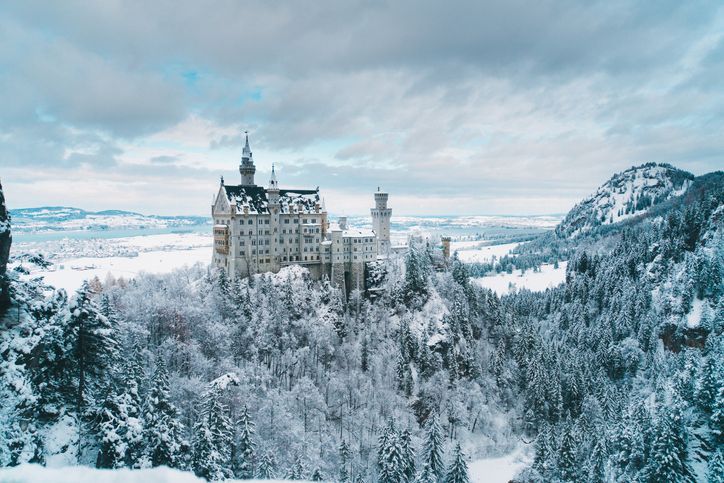 A large white castle with turrets sits on a hilltop surrounded by a dense, snow-covered forest under a cloudy winter sky.