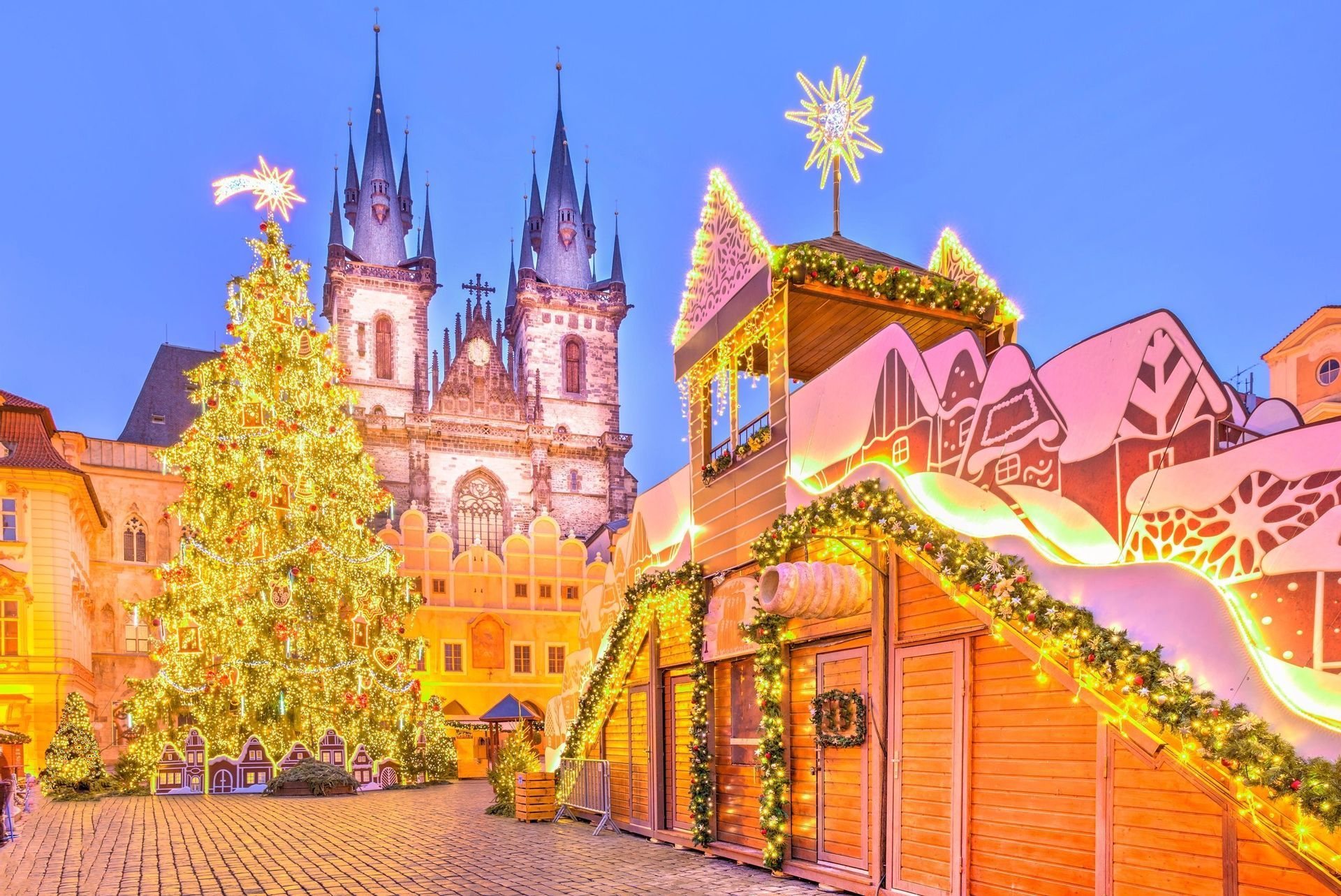 A brightly lit Christmas tree and market stalls fill a historic cobblestone square in front of a Gothic church at dusk.