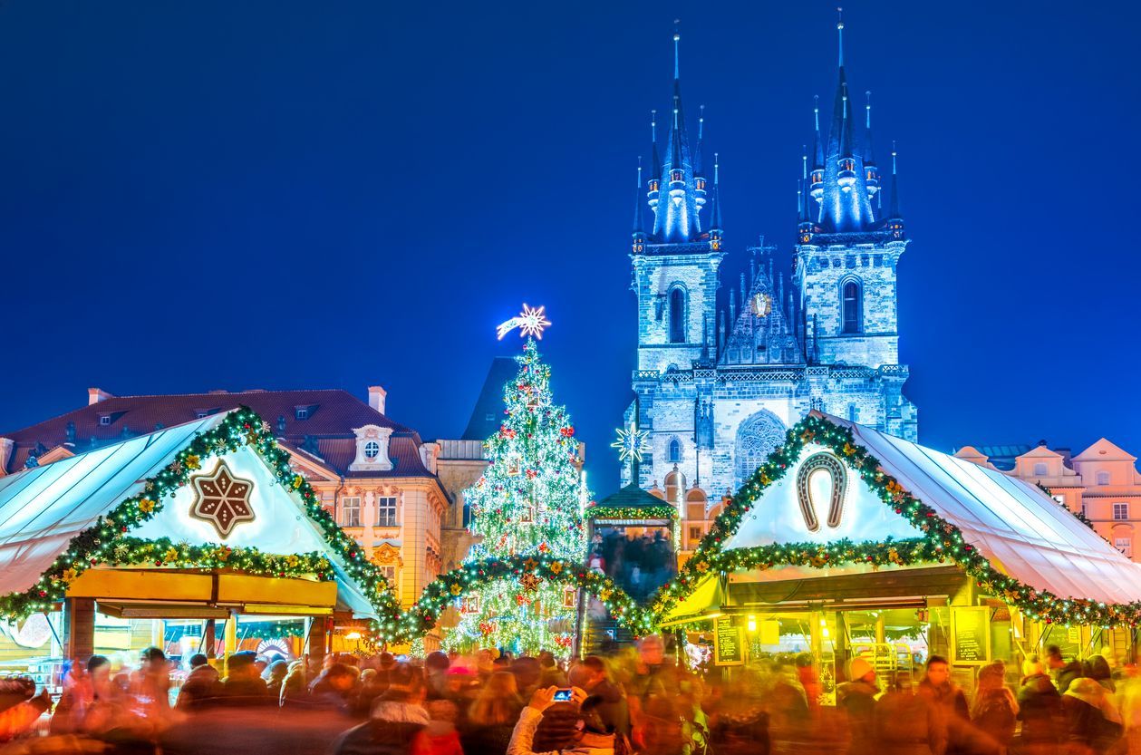 A crowded Christmas market at night with an illuminated tree and stalls in front of a large Gothic church.