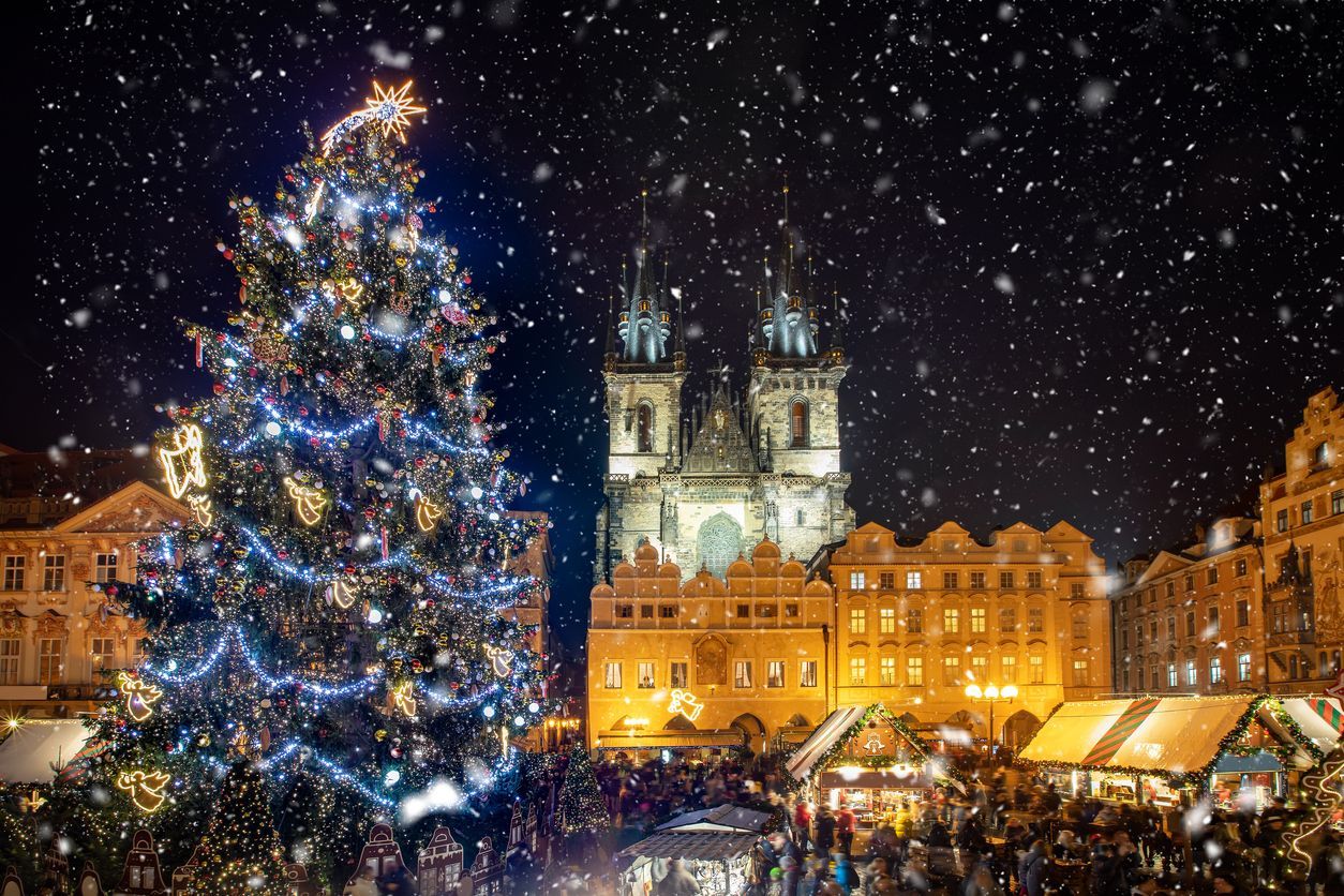 A large, illuminated Christmas tree stands in a bustling town square during a snowy Christmas market at night.