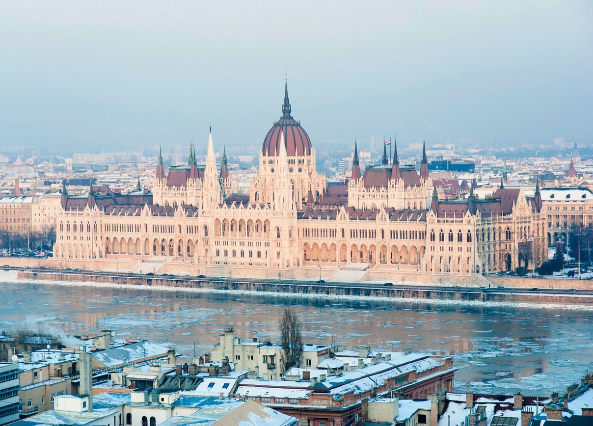 A grand, ornate building with a central dome sits on the banks of an icy river, with snow-covered city rooftops in the foreground.