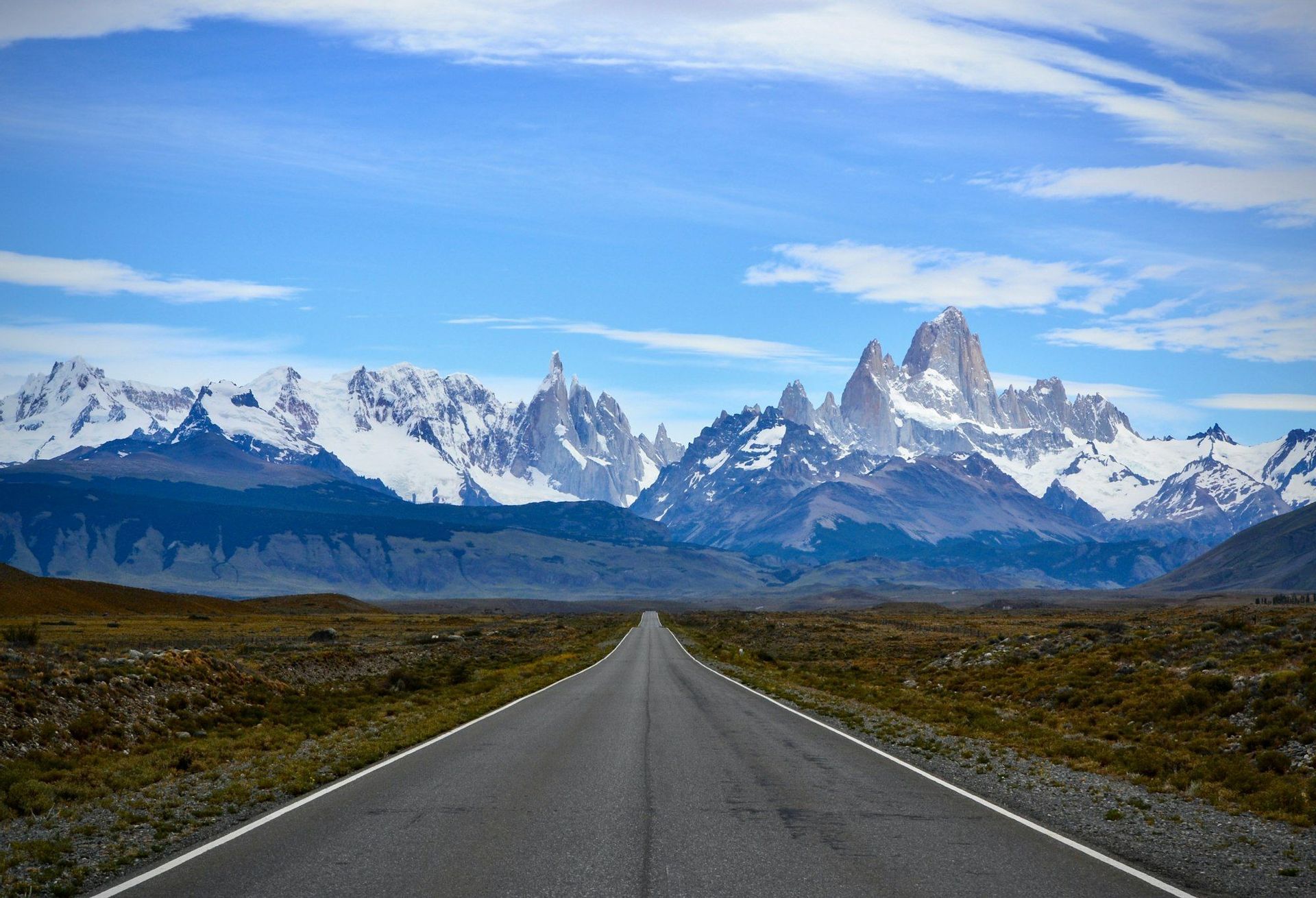 Una strada diritta e asfaltata si estende attraverso una pianura erbosa verso una catena montuosa lontana, frastagliata e coperta di neve, sotto un cielo blu.