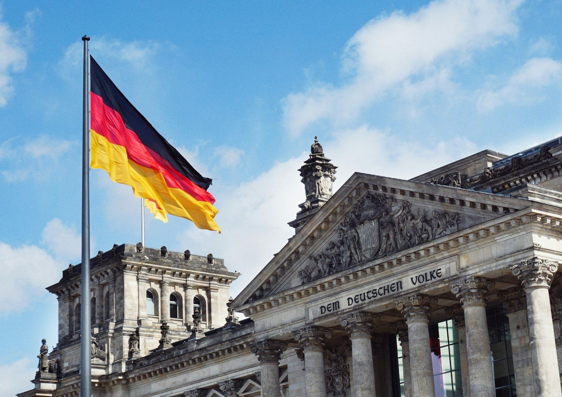 La bandera alemana ondea en un mástil junto a la fachada neoclásica del Edificio del Reichstag bajo un cielo azul parcialmente nublado.