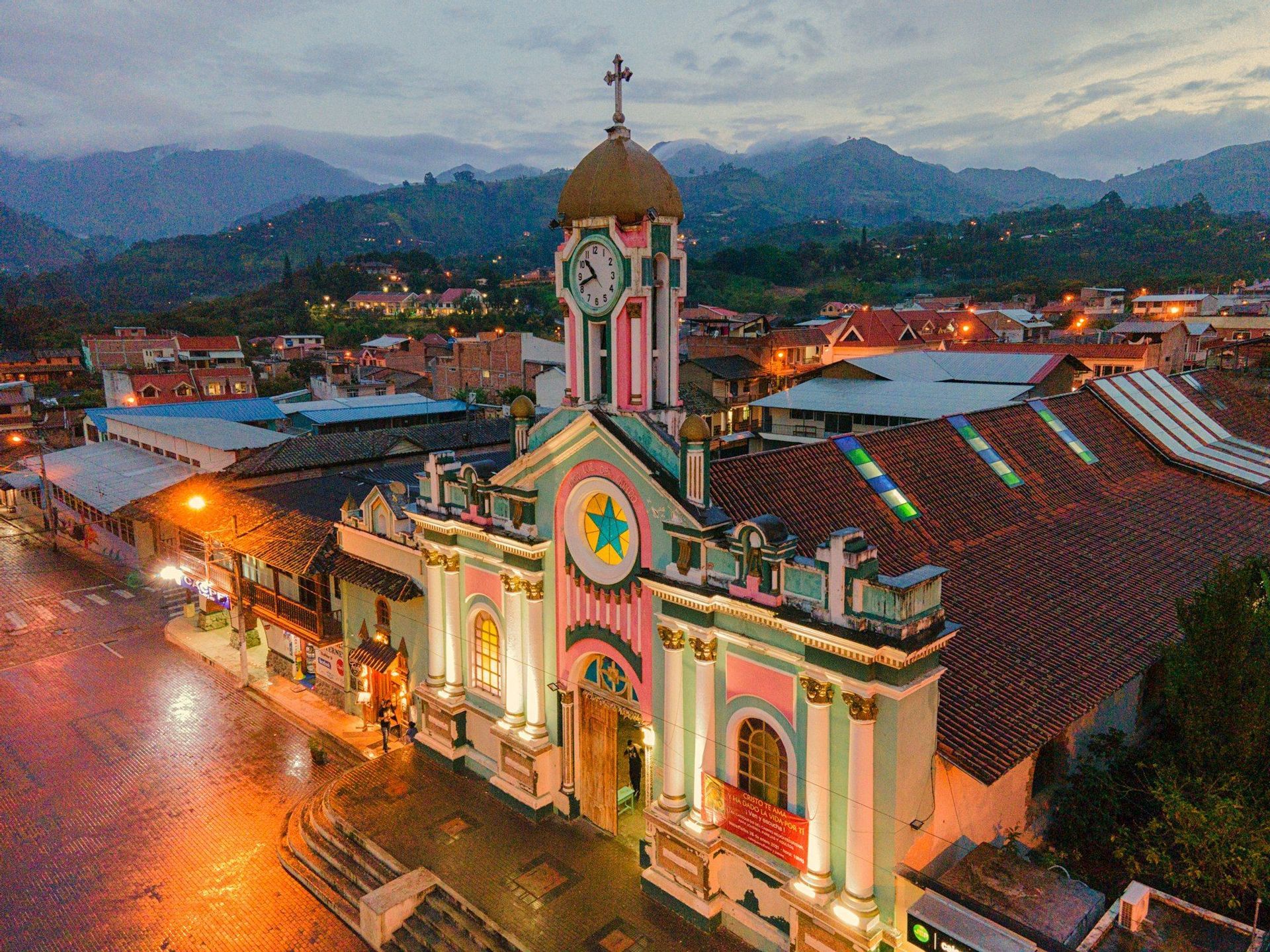 Vista aérea de una colorida iglesia rosa y verde con torre del reloj, iluminada al atardecer en un pueblo rodeado de montañas.