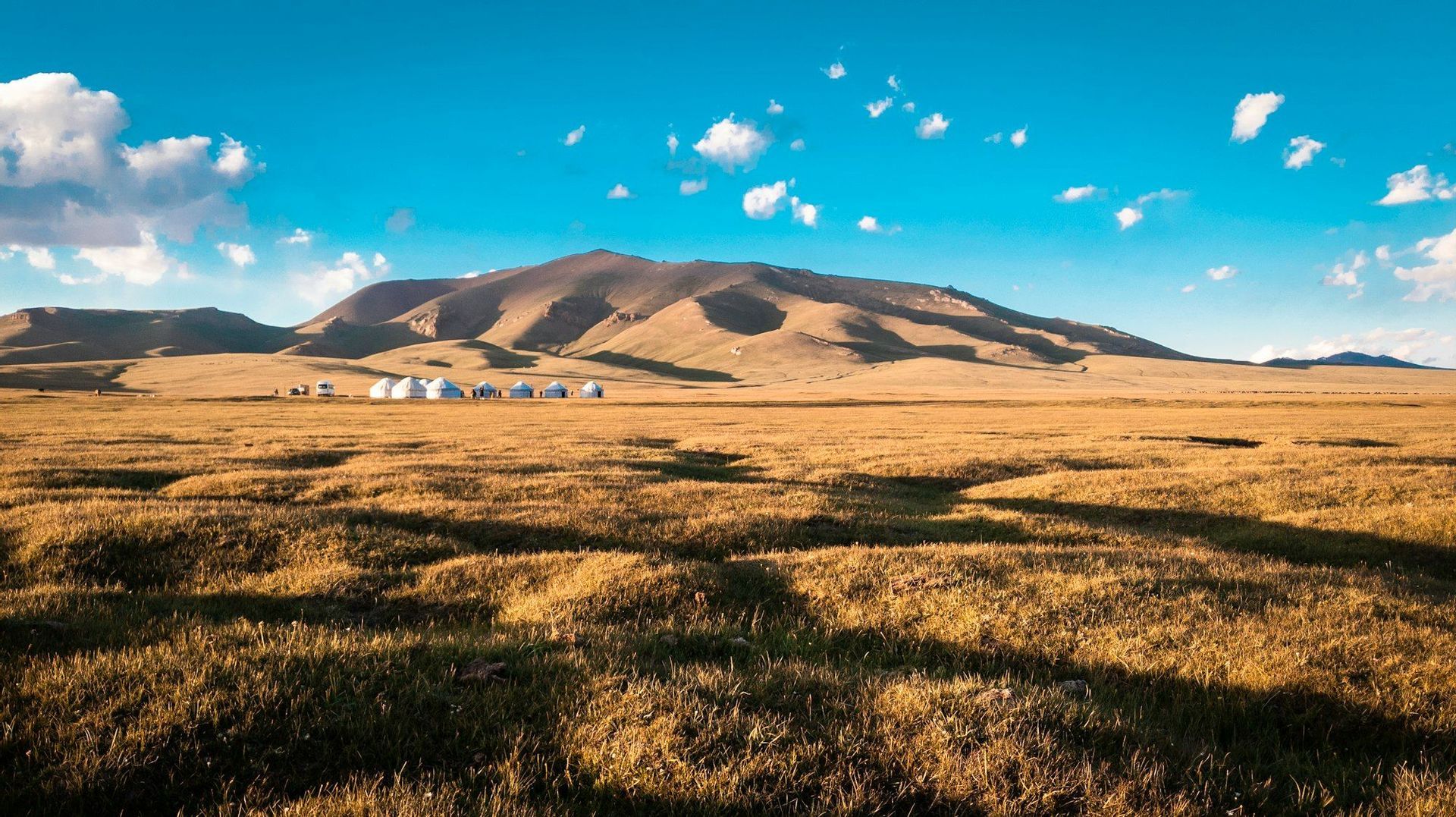 Un campamento de yurtas blancas se asienta en una vasta llanura herbosa al pie de grandes colinas onduladas, bajo un cielo azul.