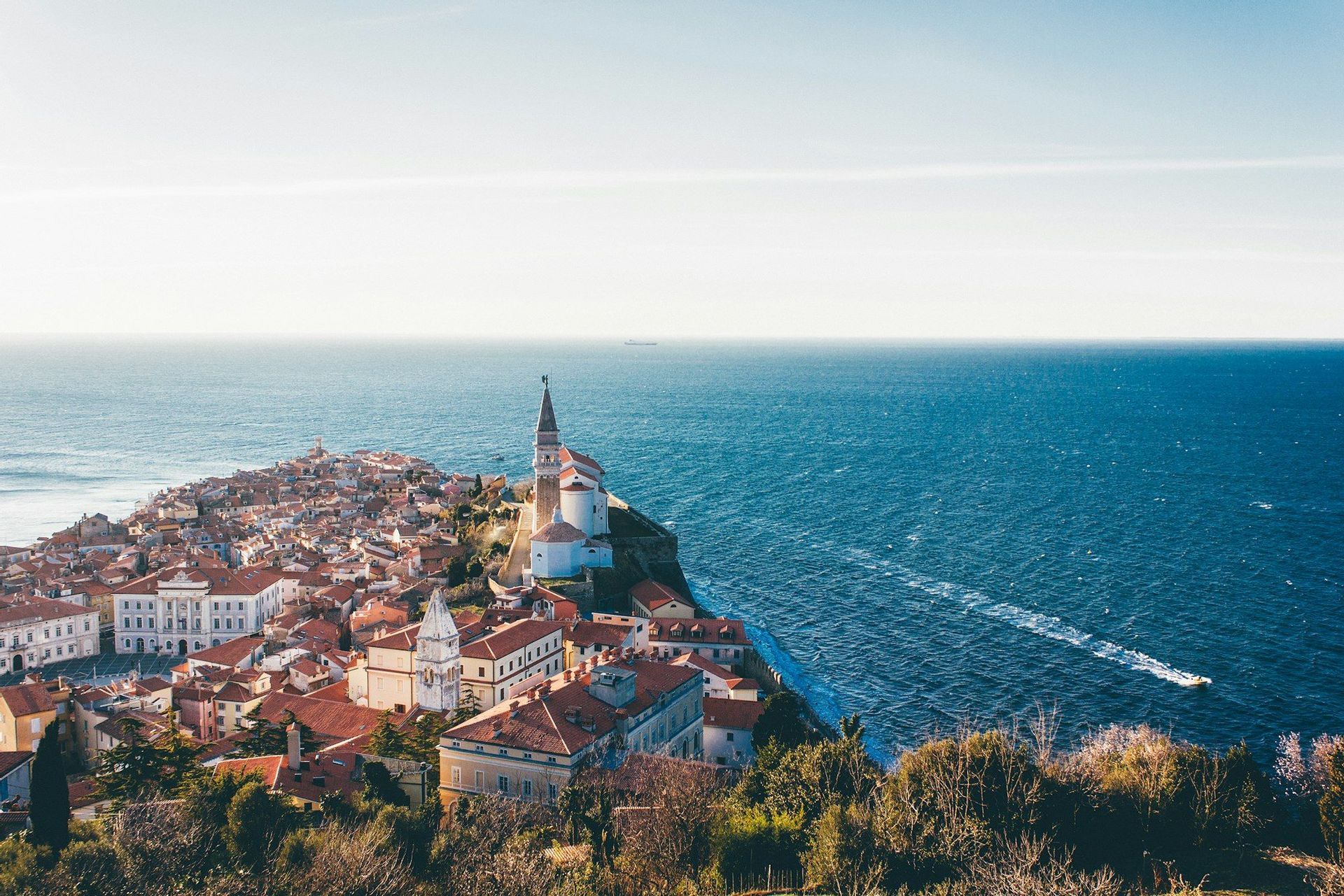 Vista aérea de un pueblo costero con tejados de teja roja y una iglesia en una península, rodeado por el mar azul profundo.