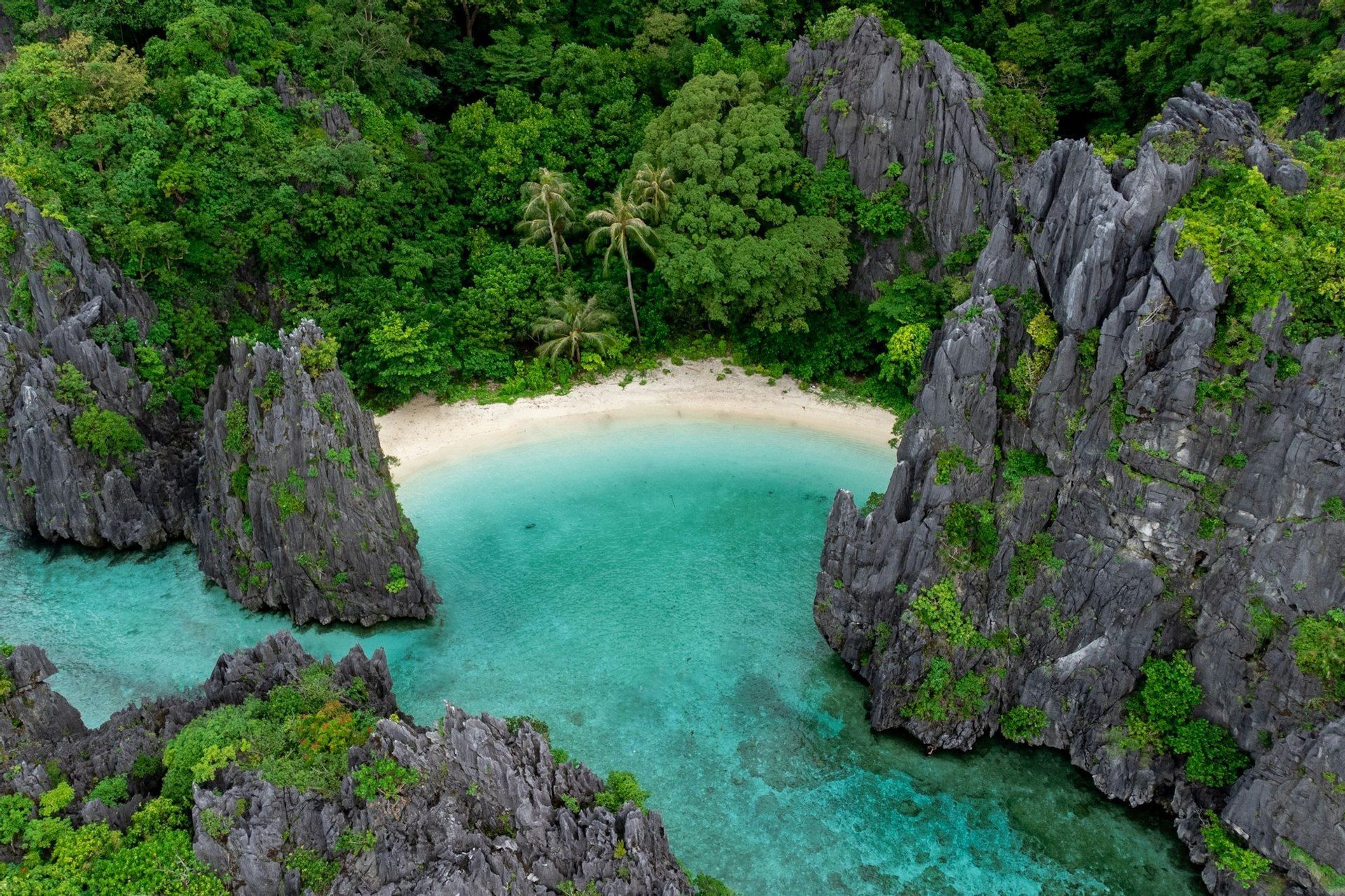 Vue aérienne d'une crique isolée avec une plage de sable blanc, entourée d'eaux turquoise, de formations rocheuses déchiquetées et d'une forêt verdoyante.