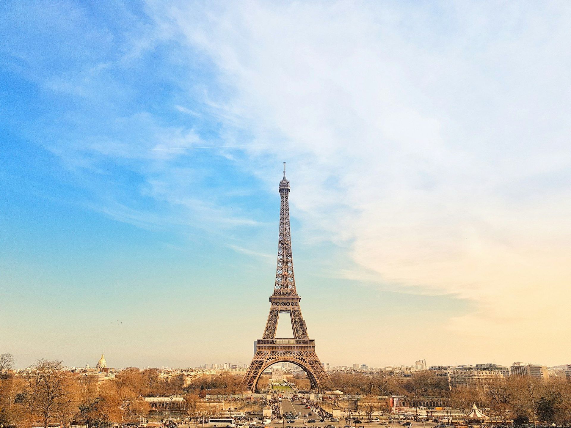 The Eiffel Tower stands against a vast blue and yellow sky, with the cityscape of Paris and streets with traffic visible at its base.