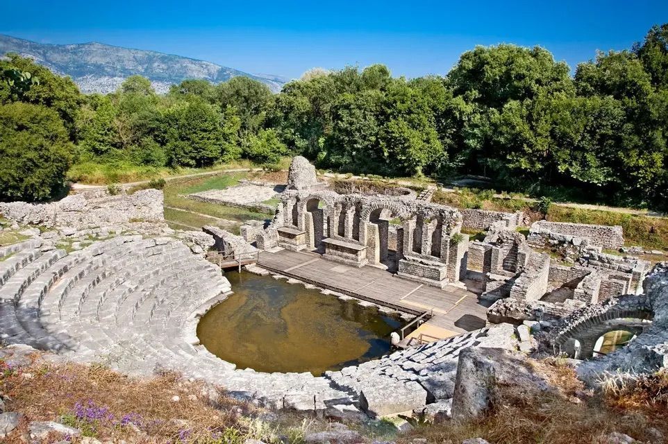 Las antiguas ruinas de un anfiteatro de piedra con un escenario inundado, rodeado de árboles verdes y montañas bajo un cielo azul claro.