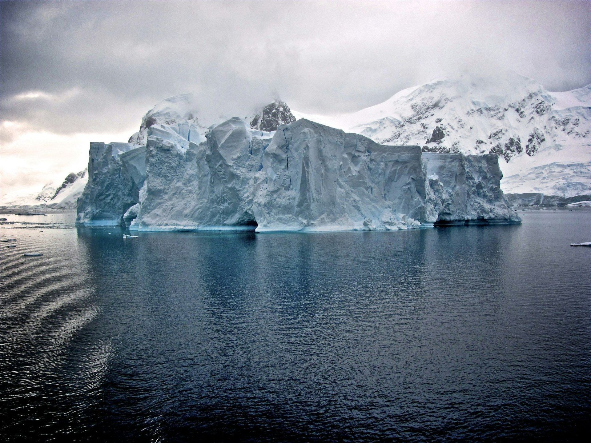 Un enorme iceberg dai bordi frastagliati galleggia in acque blu scuro, con montagne innevate sullo sfondo sotto un cielo nuvoloso.