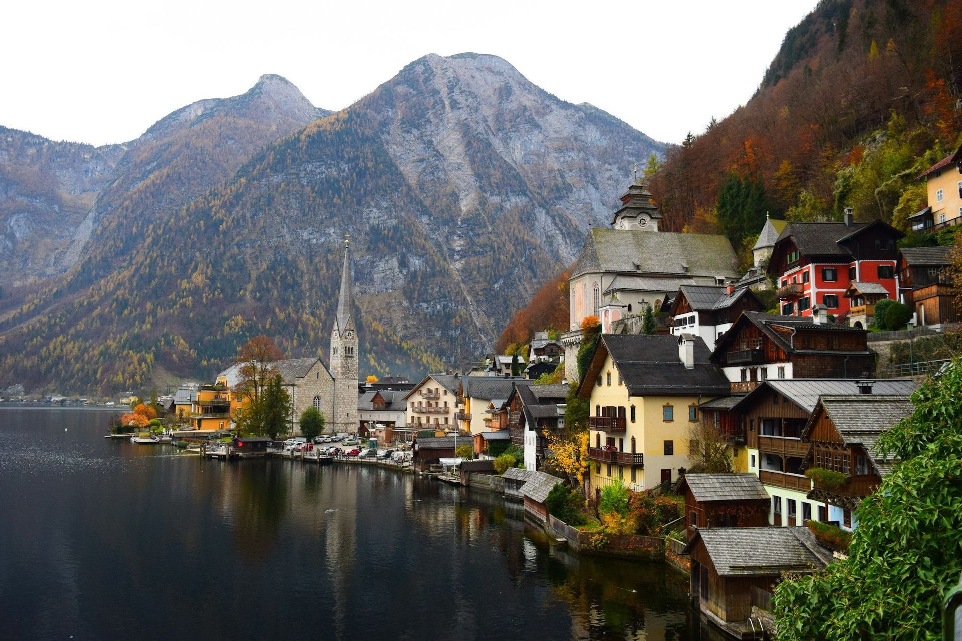 Un villaggio lacustre con un alto campanile è incastonato ai piedi di una grande montagna, con i colori autunnali sugli alberi.