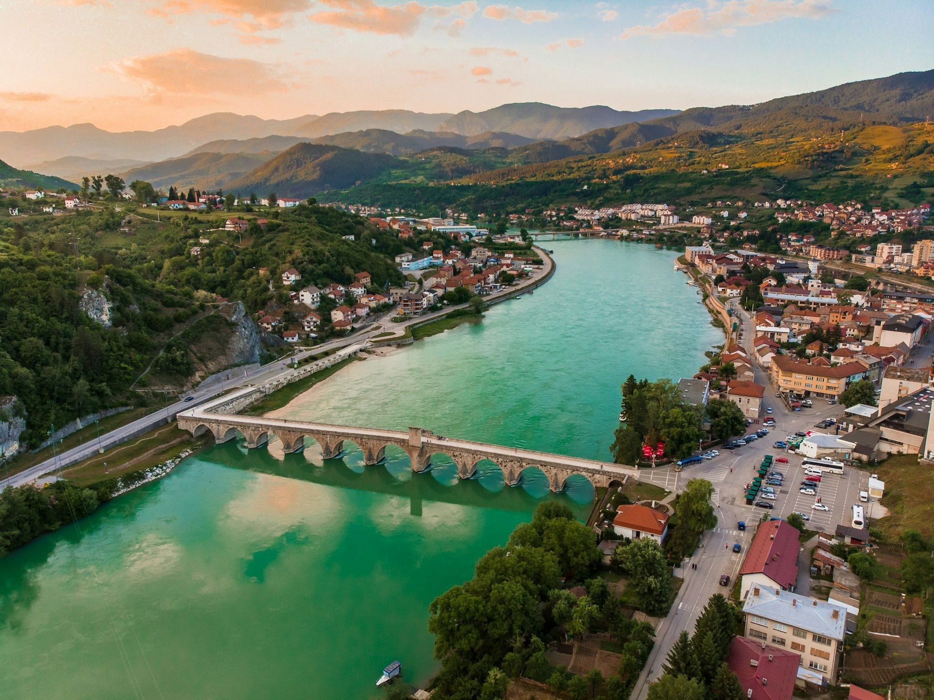 Una vista aerea di un ponte in pietra a più arcate che attraversa un ampio fiume turchese che scorre attraverso una città adagiata in una valle con montagne al tramonto.