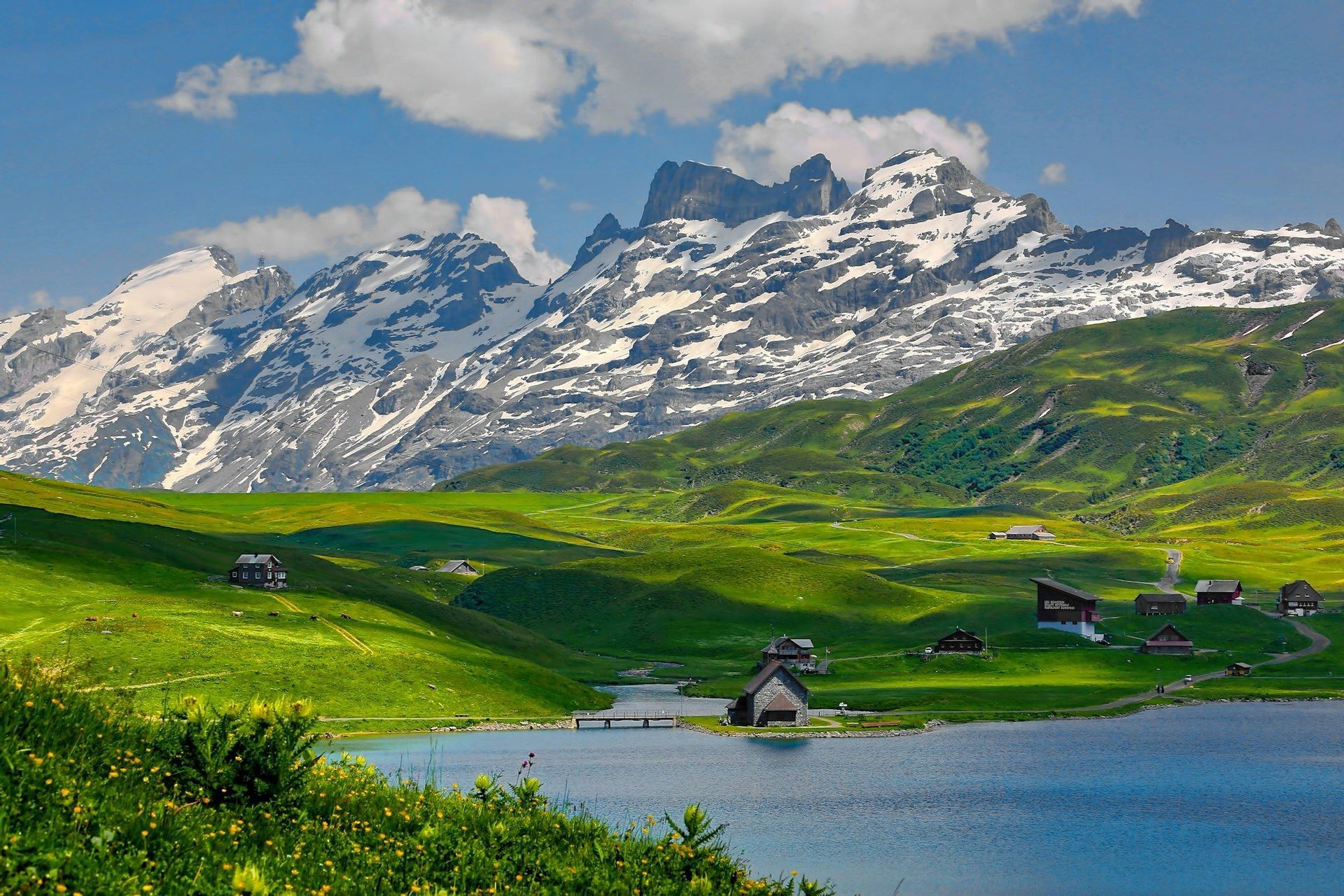 Un borgo lacustre con una cappella in pietra si trova in una valle di verdi colline sotto montagne innevate.