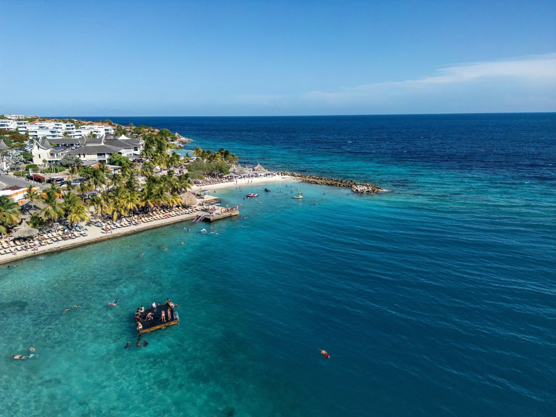 Una vista aerea di un resort tropicale con una spiaggia sabbiosa dove le persone nuotano nell'acqua turchese accanto a una piattaforma galleggiante.
