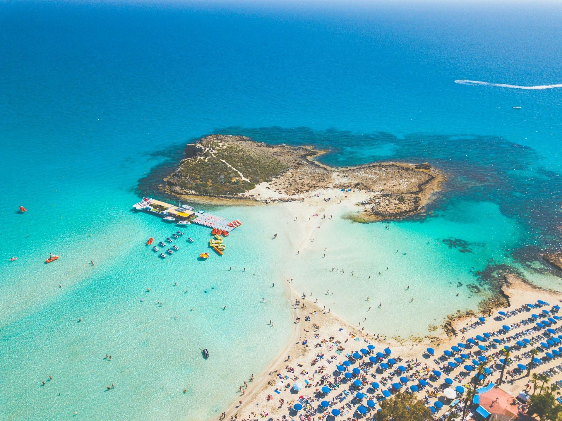 Una vista aerea di una spiaggia affollata collegata da una lingua di sabbia a una piccola isola rocciosa, con persone che nuotano in acque cristalline e turchesi.