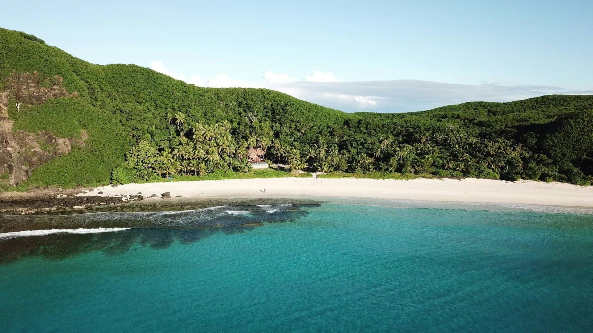 Veduta aerea di una spiaggia isolata di sabbia bianca e acque turchesi, incorniciata da lussureggianti colline verdi alberate.