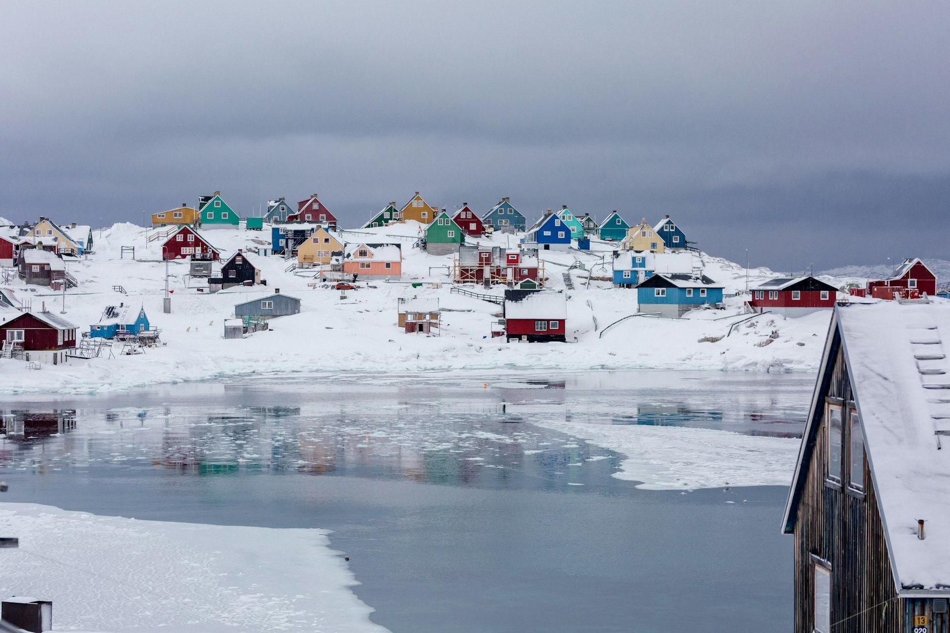 Un villaggio di case colorate si adagia su una collina innevata, vicino a uno specchio d'acqua ghiacciato, sotto un cielo nuvoloso.