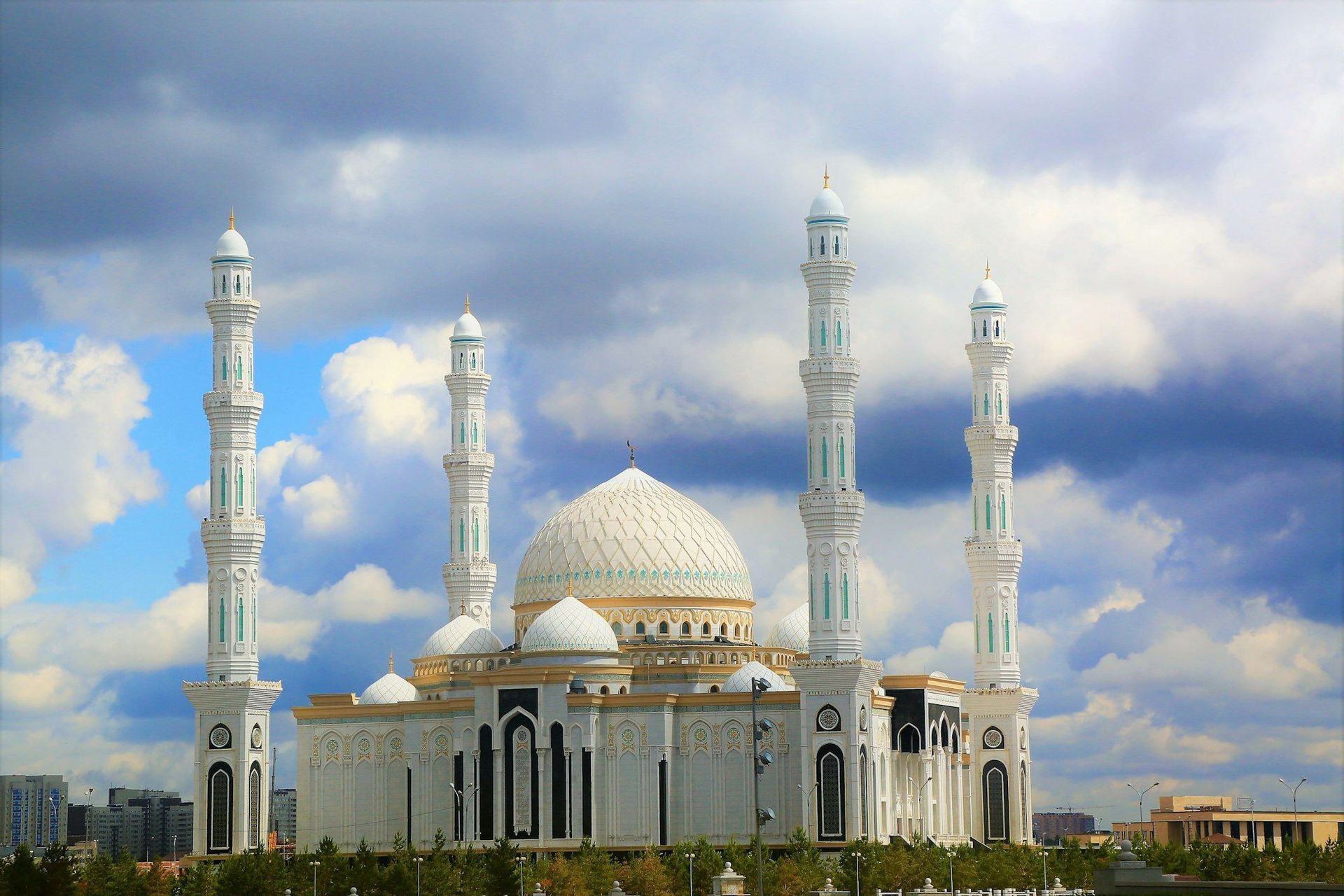 Una grande moschea bianca con cupola centrale e alti minareti, decorata con dettagli in oro e turchese, sotto un cielo azzurro e nuvoloso.