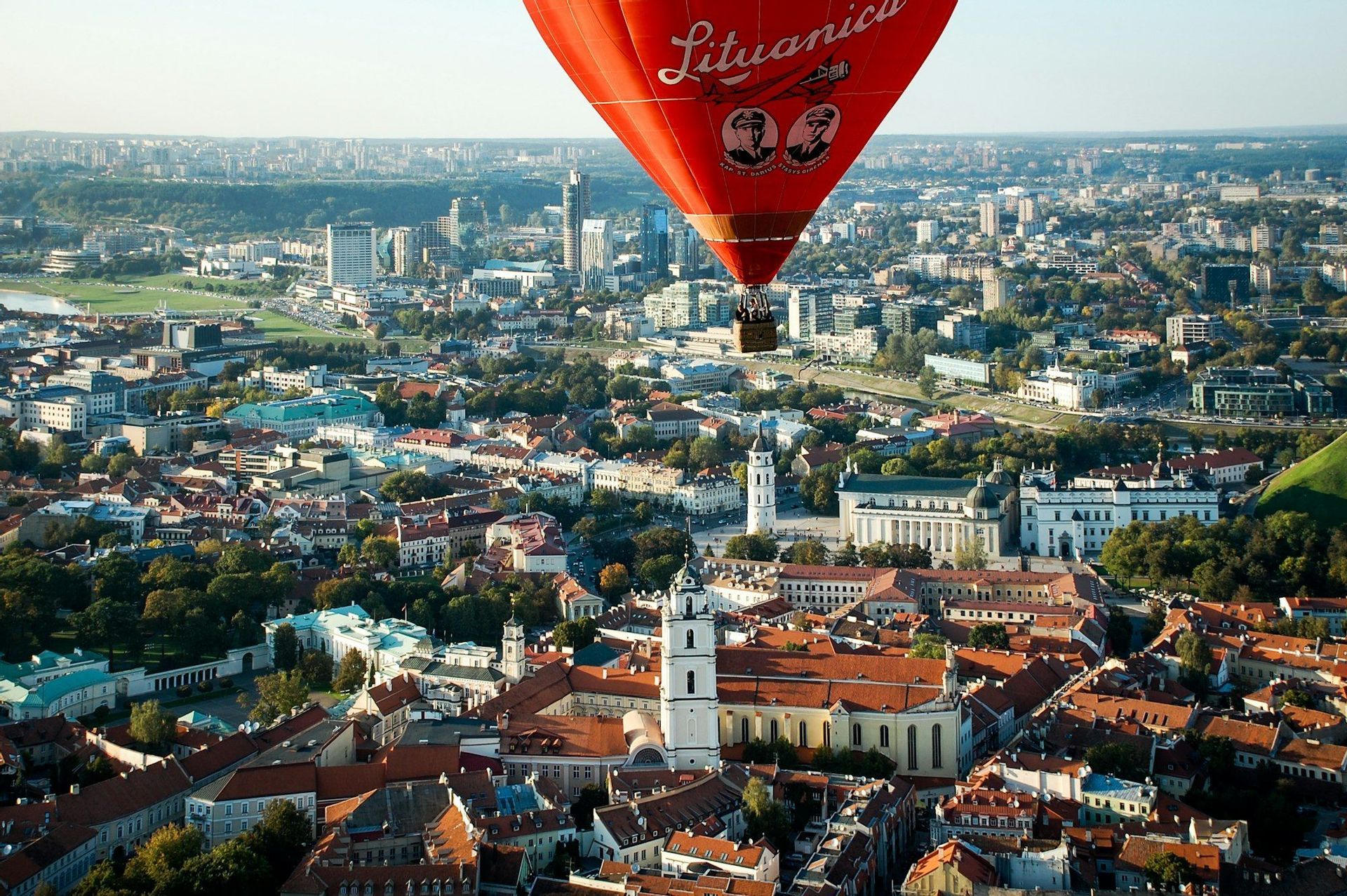 Una grande mongolfiera rossa con persone nel suo cesto fluttua sopra una città estesa con edifici dai tetti rossi e grattacieli lontani.