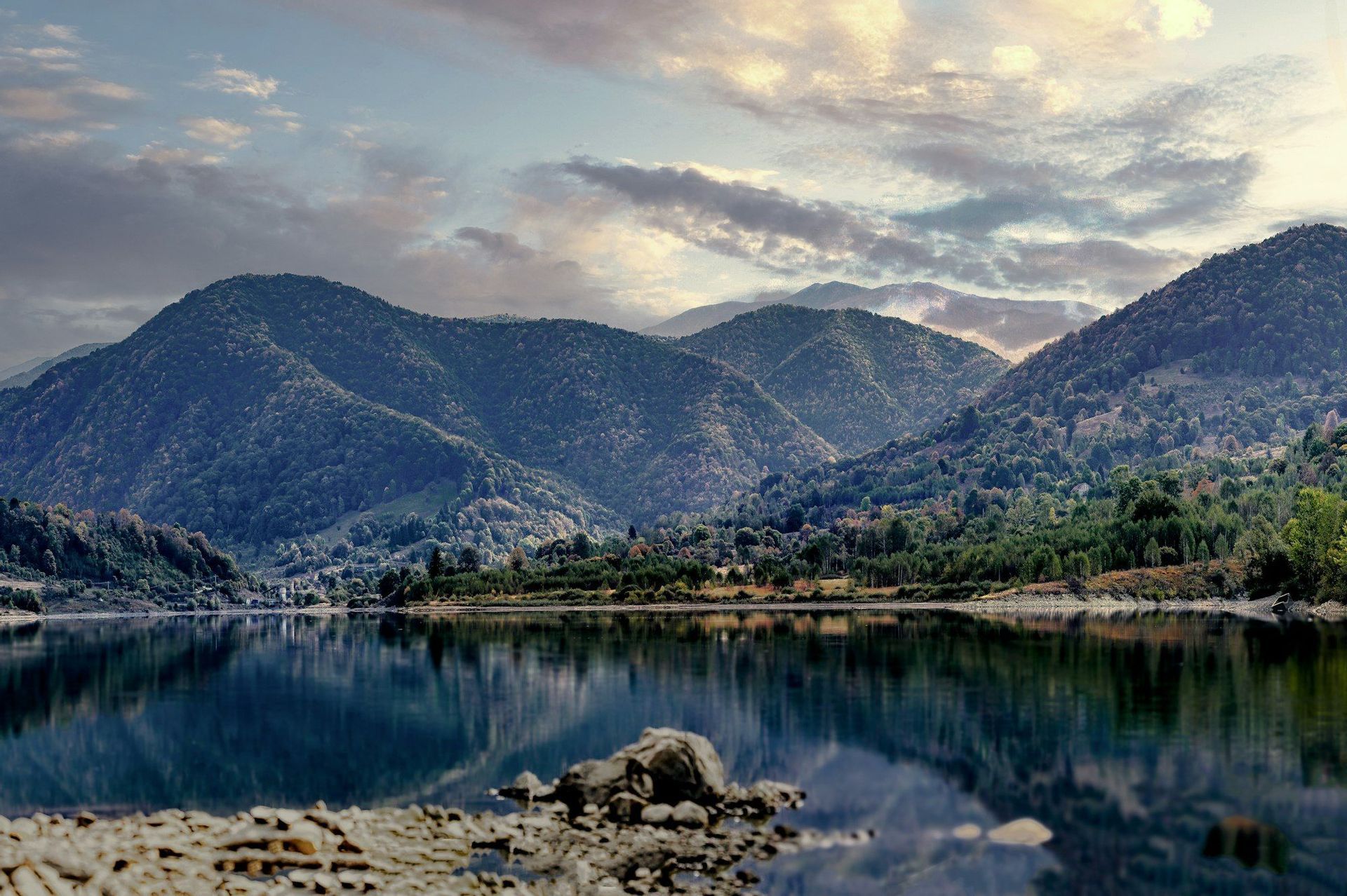 Un lago calmo riflette montagne coperte di alberi sotto un cielo nuvoloso, con una riva rocciosa in primo piano.